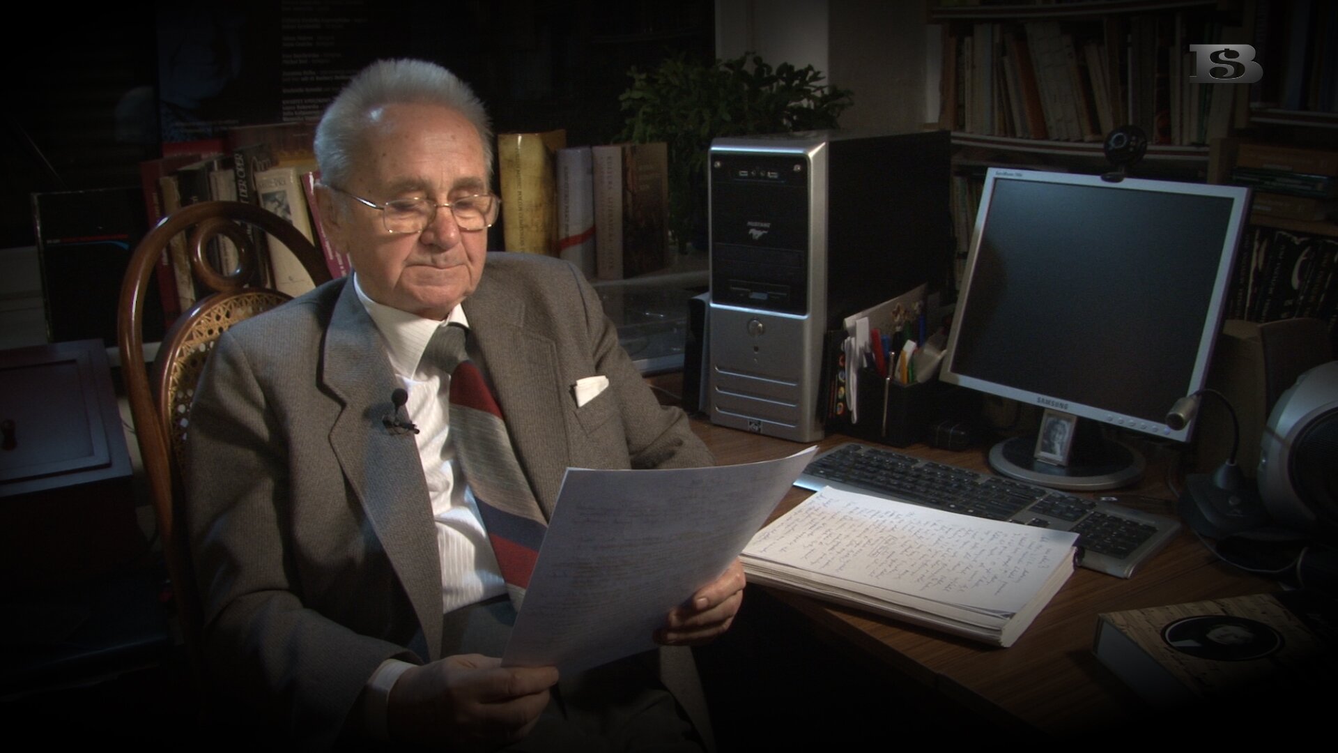 Fotografia przedstawia w biurze starszego o siwych włosach Pana siedzącego na drewnianym krześle przy biurku, który spogląda na białą kartkę papieru, którą trzyma w rękach. Człowiek ma na sobie szary garnitur, krawat w szersze paski w kolorze czerwonym, granatowym i szarym. Po prawej stronie marynarki jest przyczepiony mały, czarny mikrofon. Starszy Pan ma założone okulary do czytania. Po prawej stronie na biurku znajduje się srebrny komputer, monitor i klawiatura oraz plik białych, wypisanych papierów ułożonych jedno pod drugim. Na biurku w czarnym pojemniku, stoją również przybory do pisania, a przy papierach położona jest książka z fotografią mężczyzny. W tle na półkach znajduje się stos książek, drukarka oraz zielony kwiat. Biuro znajduje się w półmrocznym oświetleniu. 