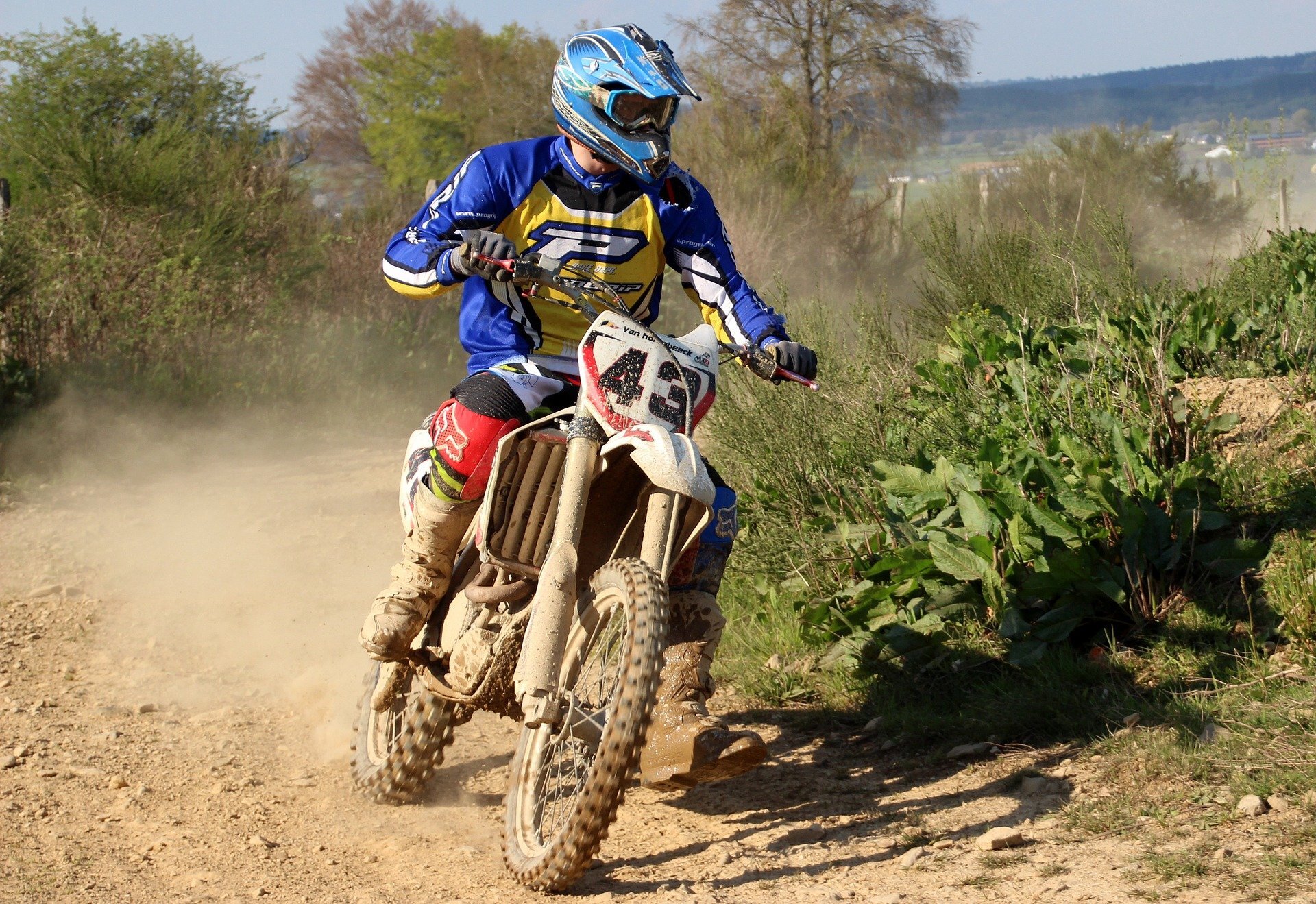 Zdjęcie ukazuje motocyklistę na motocyklu o lekkiej konstrukcji. Motocyklista porusza się w terenie po piaszczystej nawierzchni. Motocykl ma średniej grubości opony z wyraźnym bieżnikiem. Motocykl jest pokryty błotem. 