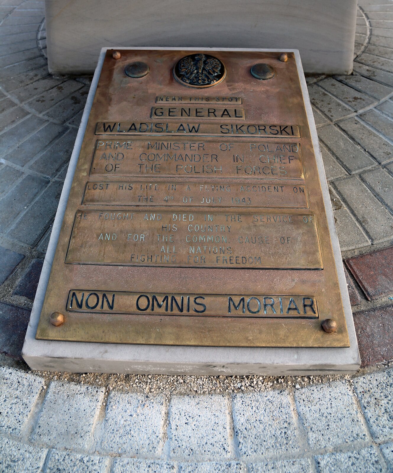 Fotografia przedstawia fragment podłoża z brukowymi płytkami, na którym znajduje się prostokątna tablica przykuta do jasnego kamienia. Tablica jest w kolorze miedzianym, są na niej czarne linijki tekstu. U góry umieszczono napis „General Wladislaw Sikorski”, a na dole „Non omnis moriar”. Pomiędzy nimi widać trzy prostokątne fragmenty z nieczytelnym tekstem. U góry widnieje orzeł z rozłożonymi skrzydłami na tle czarnego okręgu oraz dwa okręgi po jego bokach. 