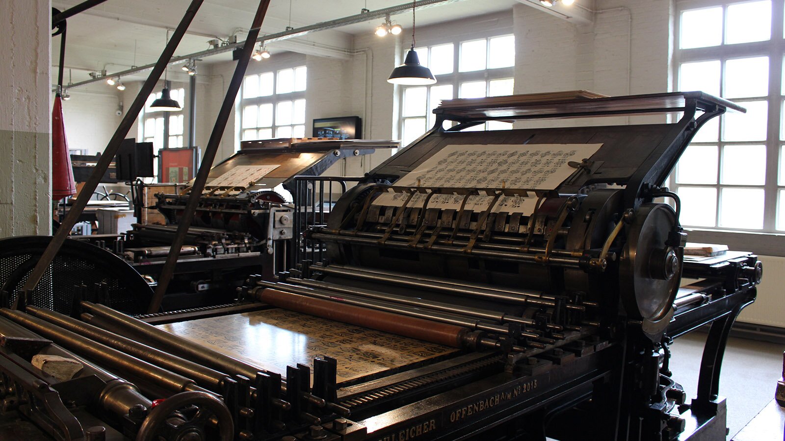 Zdjęcie przedstawia bardzo stare prasy drukarskie ustawione w dużym pomieszczeniu z wielkimi oknami. Na pierwszym planie widoczna jest żeliwna prasa z poziomym wózkiem na metalowej ramie, na którym położona jest matryca drukarska, duży cylindryczny wał, na którym przymocowana jest zadrukowana strona oraz wiele wąskich cylindrów leżących na matrycy.