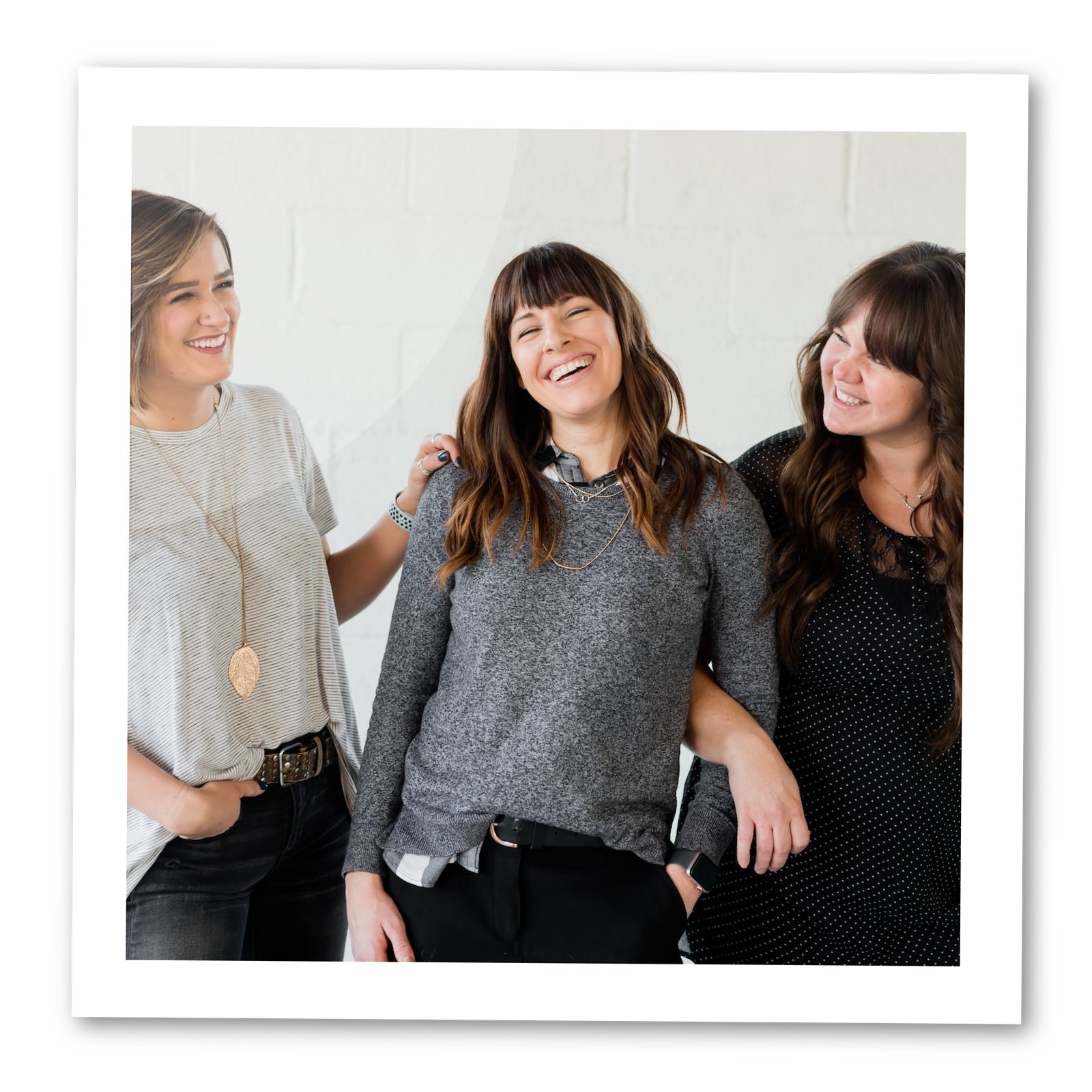 Zdjęcie przedstawia trzy kobiety w podobnym wieku, koło trzydziestki. Pierwsza z lewej ma krótkie, brązowe włosy, ubrana w jasną koszulkę i ciemne dżinsy. Ma długi, złoty wisiorek i trzyma swoją lewą dłoń na ramieniu kobiety w środku. Druga kobieta ma brązowe, falowane włosy średniej długości z grzywką. Ma na sobie szary sweter i czarne spodnie. Trzecia kobieta ma długie brązowe, falowane włosy z grzywką. Ma na sobie czarną bluzkę w białe drobne groszki. Trzyma pod rękę kobietę w środku. Pierwsza i trzecia kobieta patrzą w kierunku środkowej Pani. Wszystkie trzy szeroko się uśmiechają.