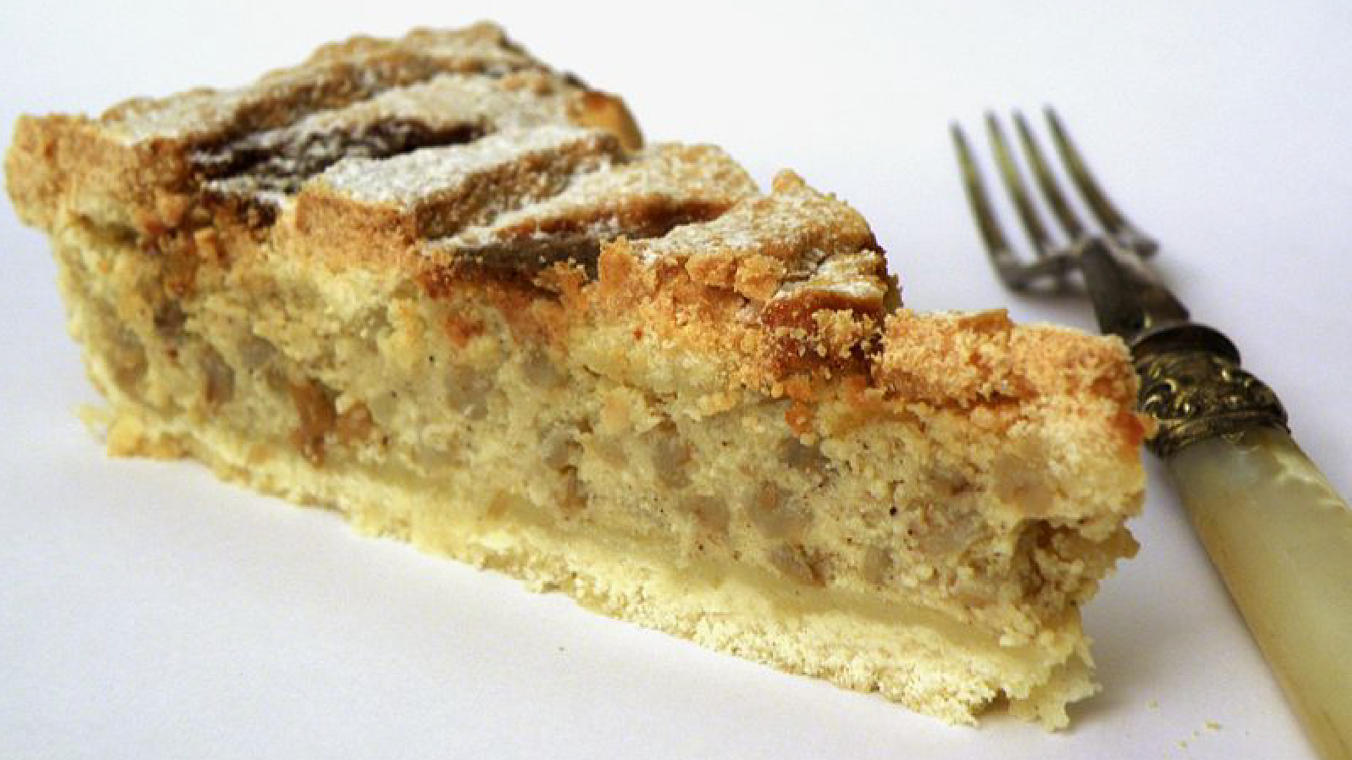 Na zdjęciu widoczny jest trójkątny kawałek placka Pastiera napoletana wypełnionego nadzieniem.  Obok ciasta leży widelec.