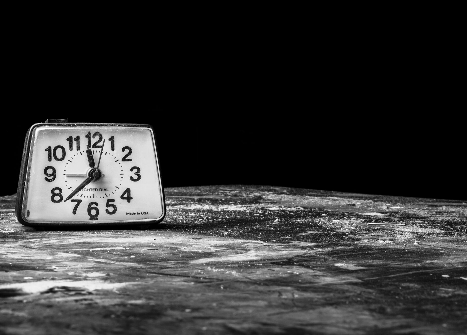 Czarno‑białe zdjęcie współczesnego wskazówkowego budzika w kształcie trapezu. Stoi na płaskiej powierzchni, która przypomina lód albo skałę, ma niejednolity kolor, gdzieniegdzie widać rozsypane drobinki. Tło jest czarne.  Budzik ma ciemną delikatną obudowę i białą tarczę. Cyfry od 1 do 12 są duże i wyraźne, minuty i godziny zaznaczone są małymi kreseczkami. Wskazówki pokazują godzinę 11.39,  budzik nastawiony jest na godzinę 9.00. 