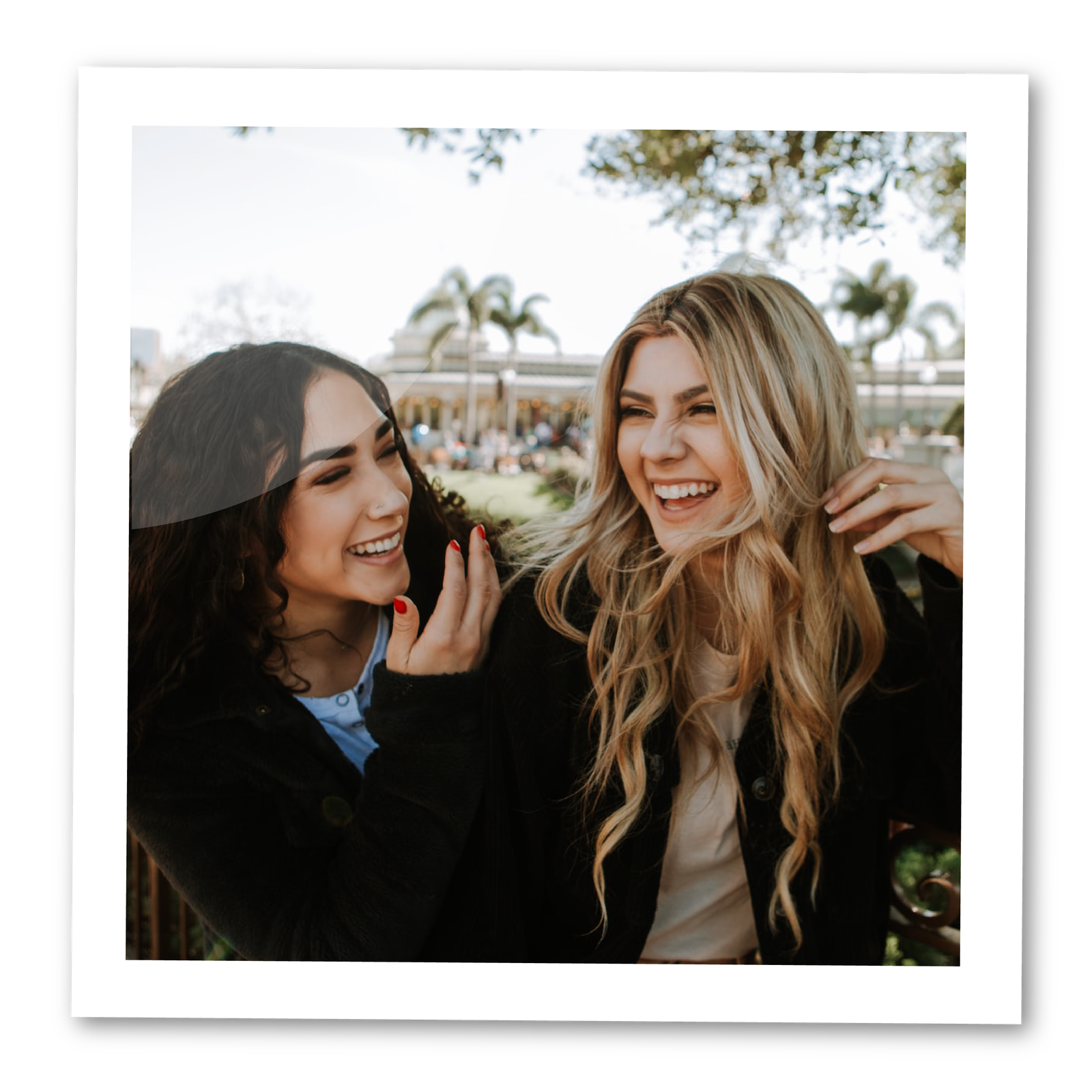 Zdjęcie przedstawia dwie młode kobiety. Obie szeroko się uśmiechają. Kobieta z lewej patrzy w kierunku tej z prawej, ma ciemne, kręcone włosy do ramion i ubrana jest w czarny sweter spod którego wystaje niebieska bluzka. Trzyma prawą rękę przy twarzy, ma paznokcie pomalowane na czerwono. Kobieta z prawej strony ma długie blond włosy, ma na sobie czarny płaszcz i beżową bluzkę. Lewą rękę trzyma trzy włosach bawiąc się nimi.