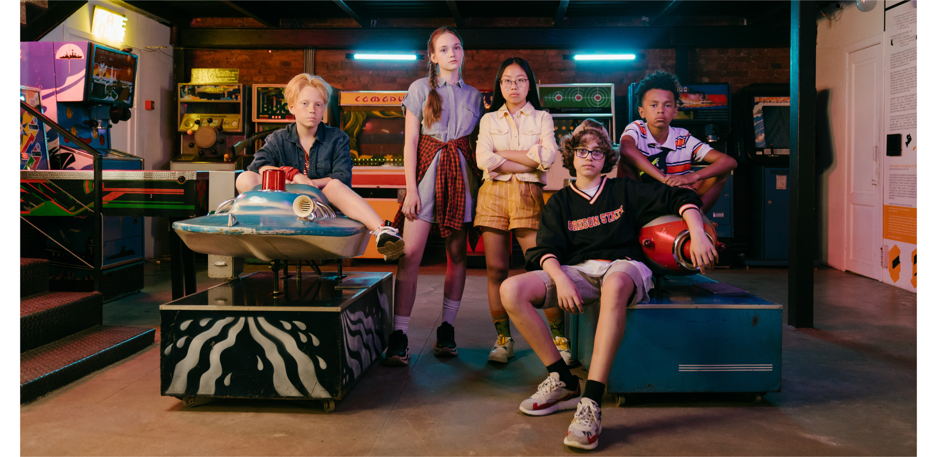 Zdjęcie przedstawia pięcioro nastolatków w salonie gier. Pierwszy z lewej na urządzeniu w salonie siedzi chłopak z dość krótkimi, rudymi włosami. W środkowej części zdjęcia stoją dwie dziewczyny. Dziewczyna z lewej jest szczupła, wysoka, ma długie włosy zaczesane w warkocz. Dziewczyna po prawej jest niższa, ma długie, proste czarne włosy. Nosi okulary. Po prawej stronie na urządzeniu siedzi dwóch chłopaków. Ten z lewej ma dłuższe kręcone włosy, nosi okulary i ma na głowie czapkę założoną daszkiem do tyłu. Chłopak po prawej ma ciemną karnację i krótkie kręcone czarne włosy.