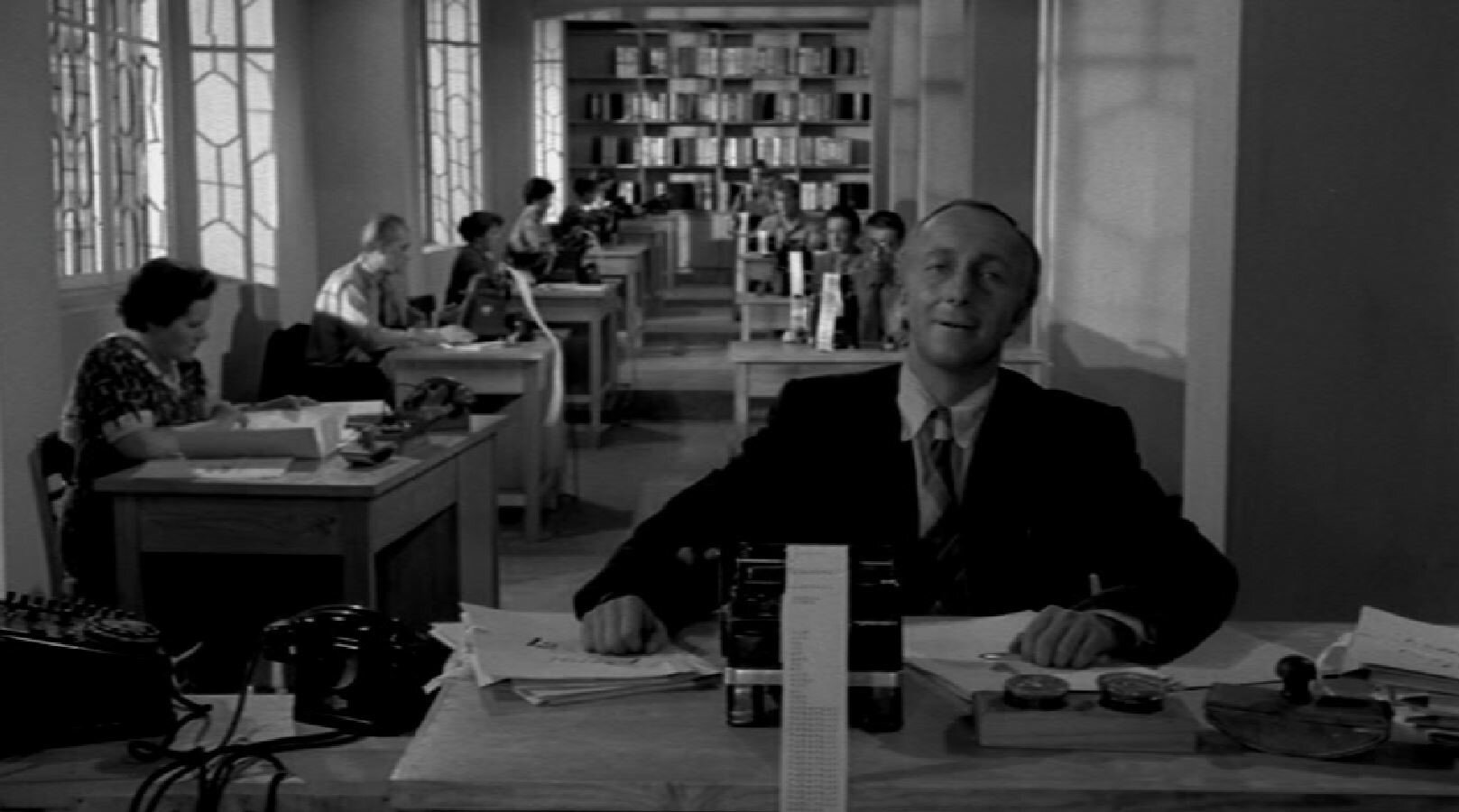 Zdjęcie przedstawia duże pomieszczenie, w którym znajdują się liczne biurka. Siedzą przy nich urzędniczki i urzędnicy. Na pierwszym planie ukazany jest także główny bohater siedzący przy biurku.