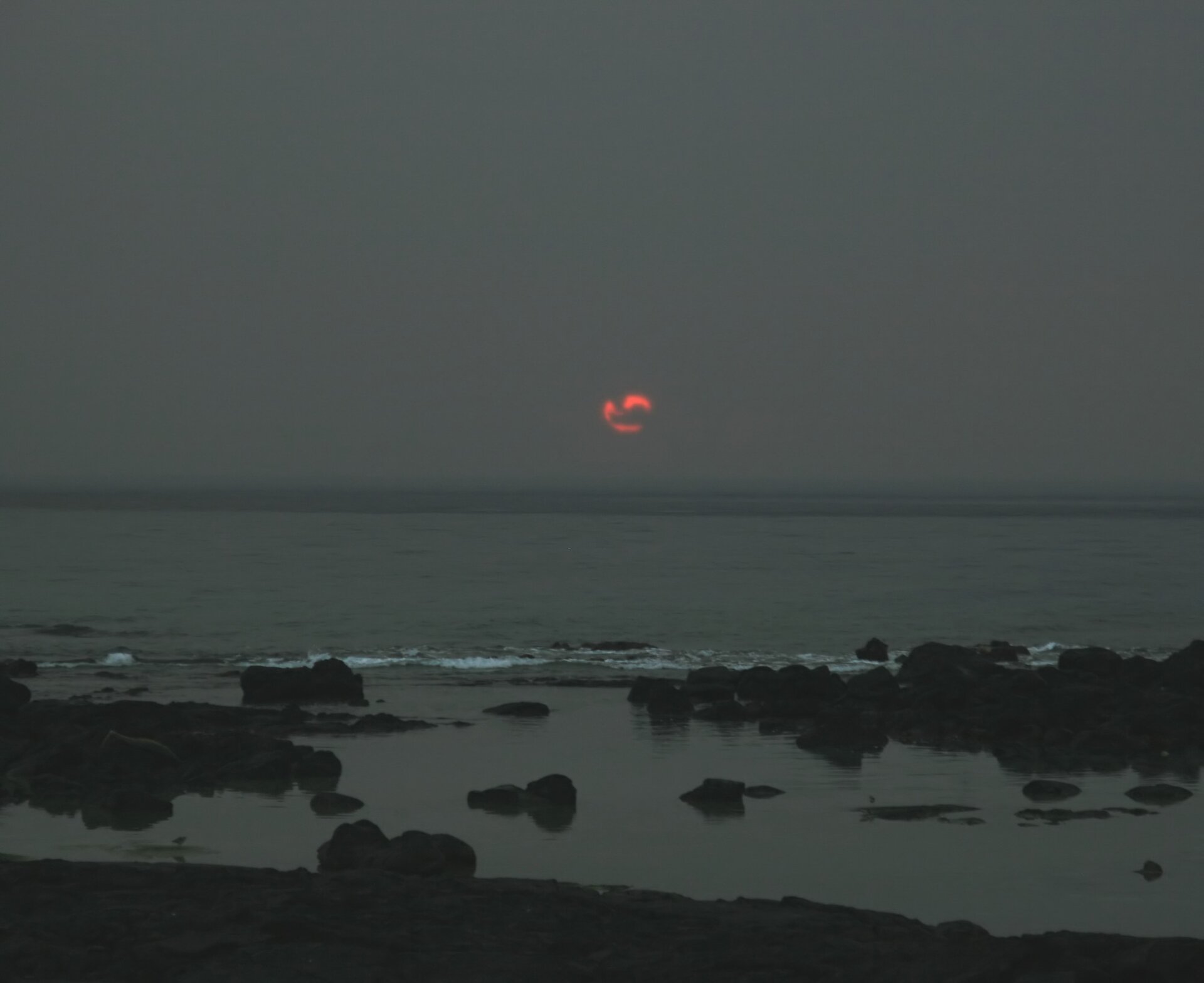 Zdjęcie przedstawia ciemne, szare niebo nad morzem. Na horyzoncie jest czerwone, zachodzące słońce.  