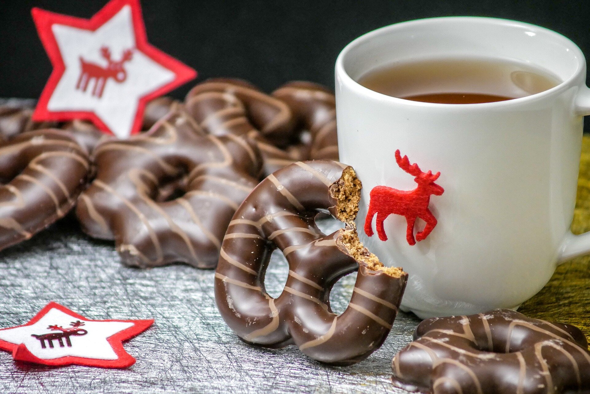 Na zdjęciu pierniki w czekoladzie i kubek z herbatą. Na kubku jest ozdobny renifer. Jeden z pierników jest nadgryziony i oparty o kubek z herbatą. Przy piernikach leżą dwie gwiazdki wycięte z cienkiego filcu. Na środku gwiazdek są wizerunki reniferów.  
