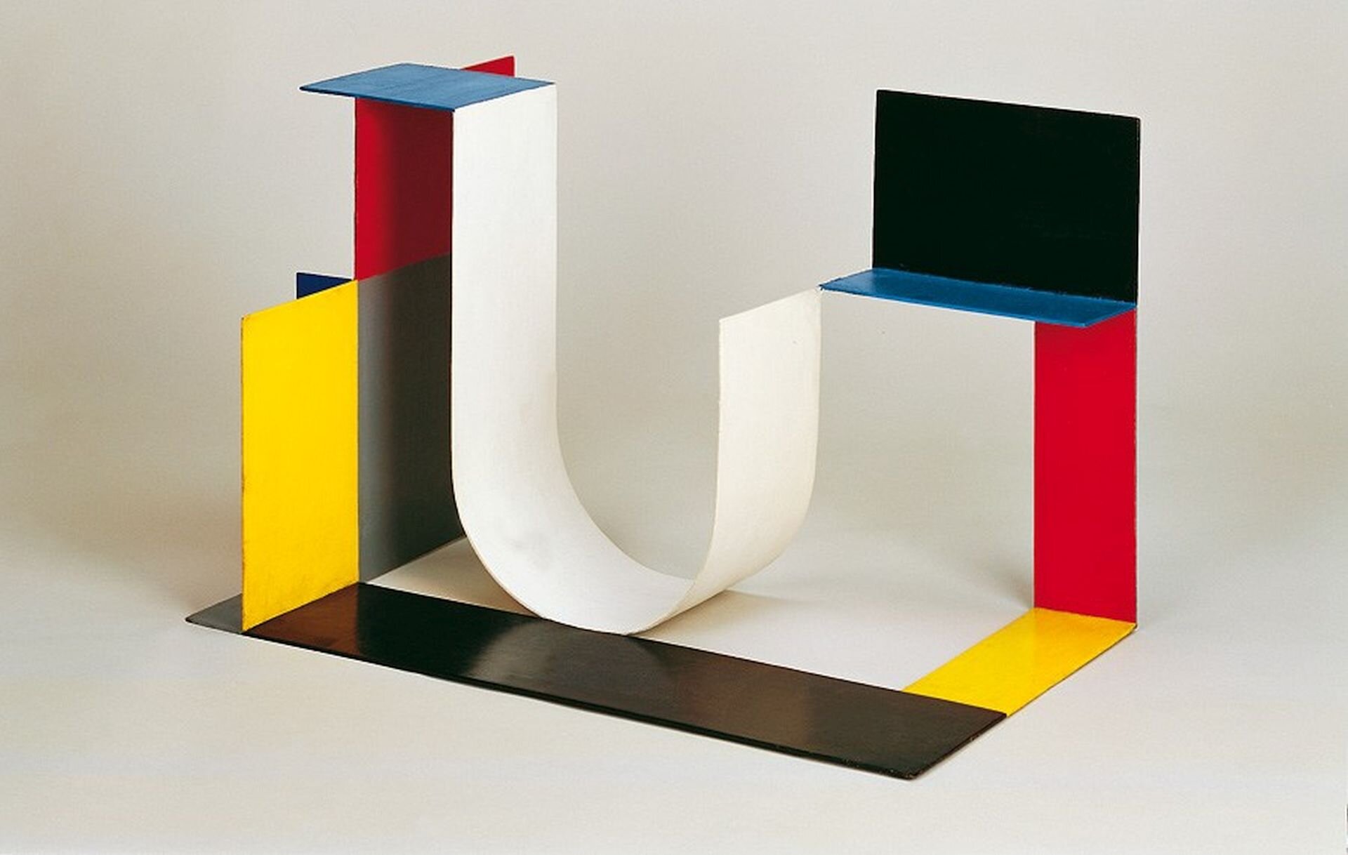 Ilustracja przedstawia rzeźbę Katarzyny Kobro pt. „Kompozycja przestrzenna 4”. Rzeźba w kształcie litery U stworzona z różnych figur, w kolorze czarnym, niebieskim, szarym, żółtym, białym, czerwonym.