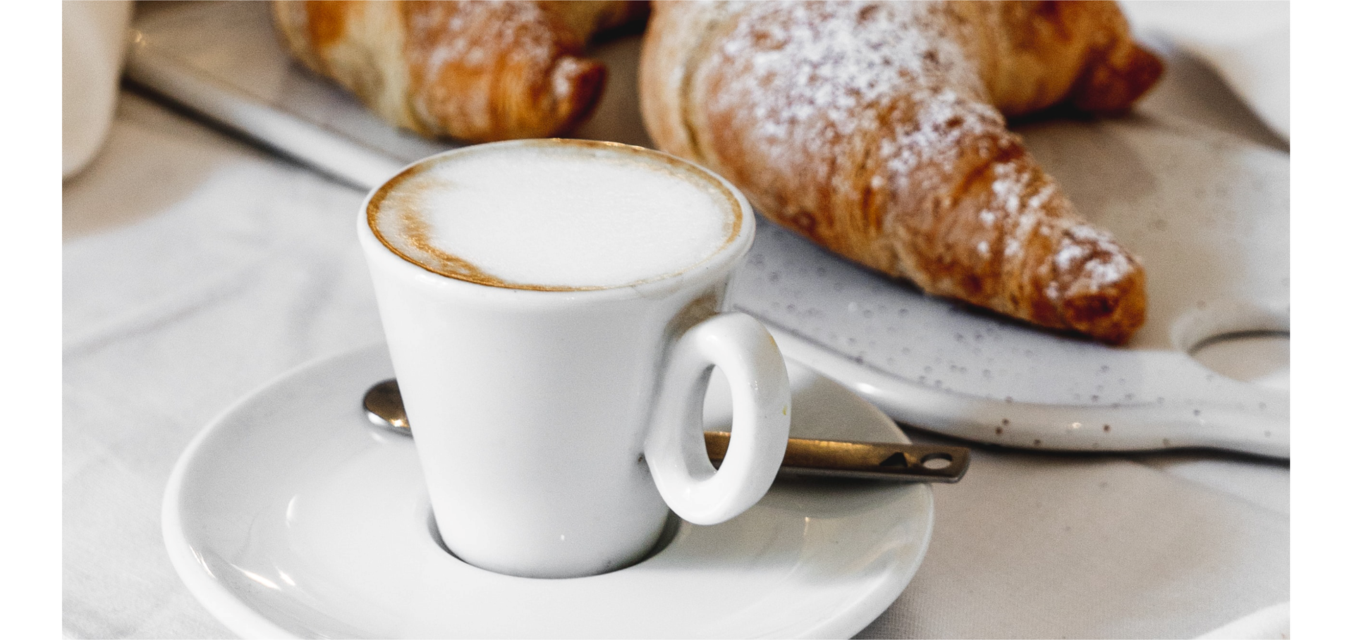 Zdjęcie przedstawia włoskie śniadanie. Na pierwszym planie znajduje się filiżanka ze spodkiem. W filiżance znajduje się kawa z mleczną pianką. Na dalszym planie widoczne są rogaliki posypane cukrem pudrem.