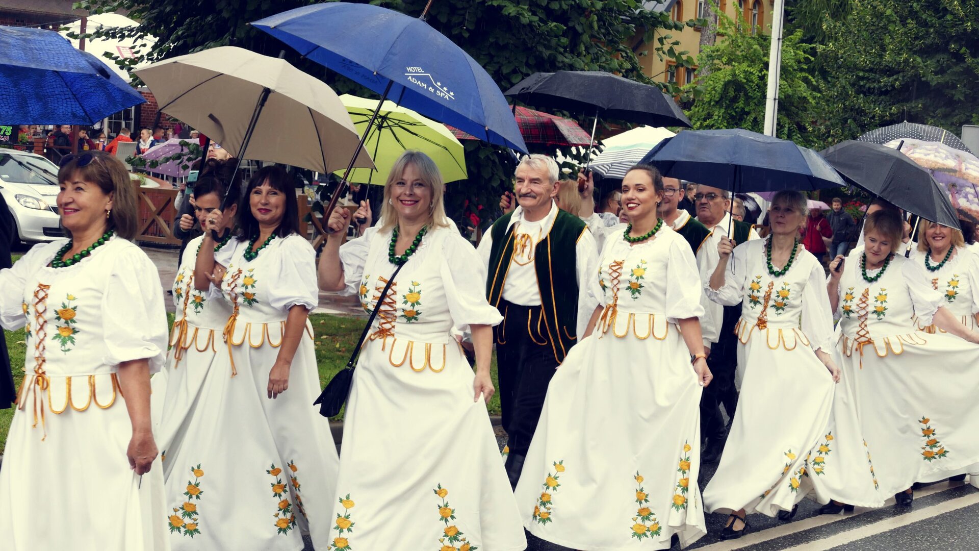 Na ilustracji znajduje się grupa kobiet idących ulicą ubranych w takie same suknie. Suknie są białe. Na sukniach znajdują się elementy żółtych kwiatów z listkami. Wszystkie kobiety mają na sobie zielone korale. Kobiety trzymają w rękach parasole. Między kobietami znajdują się mężczyźni ubrani w białe koszule, zielone spodnie oraz zielone kamizelki. W tle zdjęcia znajdują się drzewa oraz tłum ludzi. 