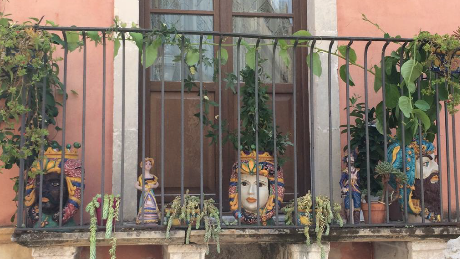 Zdjęcie przedstawia balkon w sycylijskim mieście Taorminie. Widoczne są pnące rośliny okalające barierę balkonu. Niektóre z nich posadzone są w wazach zwanymi głowami Maurów. Widoczne są również drewniane drzwi balkonowe oraz fragment muru budynku.