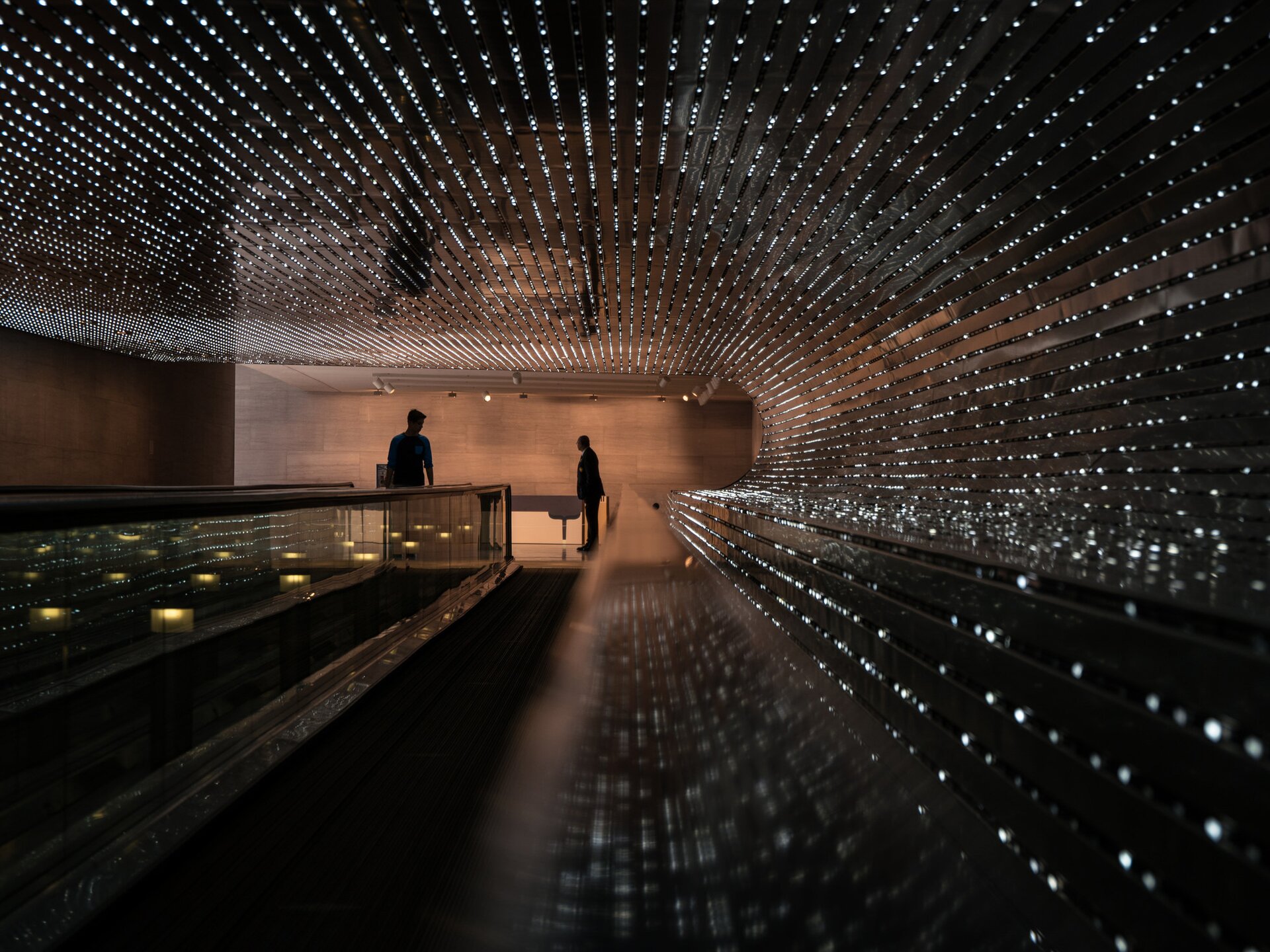 Fotografia barwna. Instalacja świetlna amerykańskiego artysty Leo Villareala w 61-metrowym tunelu Narodowej Galerii Sztuki w Waszyngtonie. Instalacja składa się z 41 000 diod Led zamontowanych w równych odstępach na ścianie i na suficie, wzdłuż ruchomego chodnika. W głębi tunelu, przed chodnikiem, sylwetki dwóch mężczyzn.