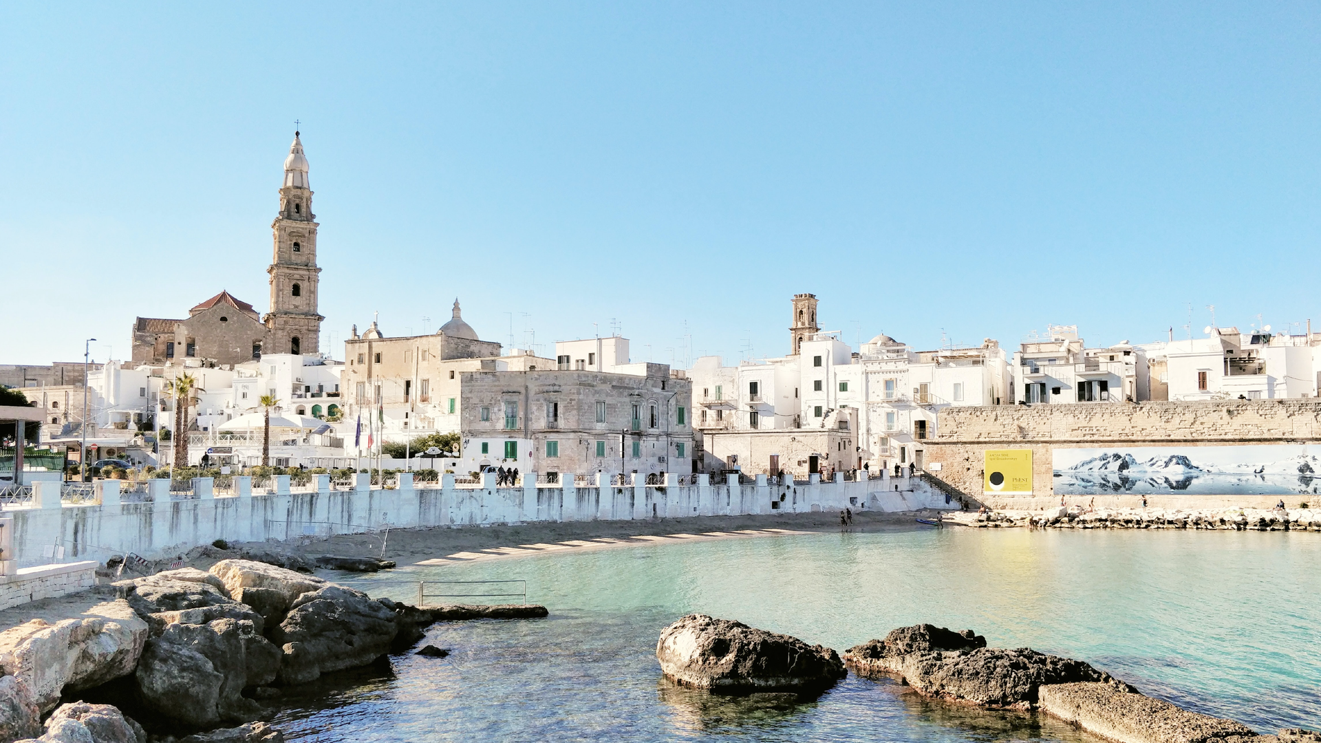 Na zdjęciu pokazane jest stare, włoskie miasto znajdujące się nad wodą. Przy plaży wznosi się promenada. Brzegiem morza chodzą ludzie. Ponad panoramą miasta wznosi się wieża kościoła. Brzeg bliżej obiektywu jest bardzo skalisty.