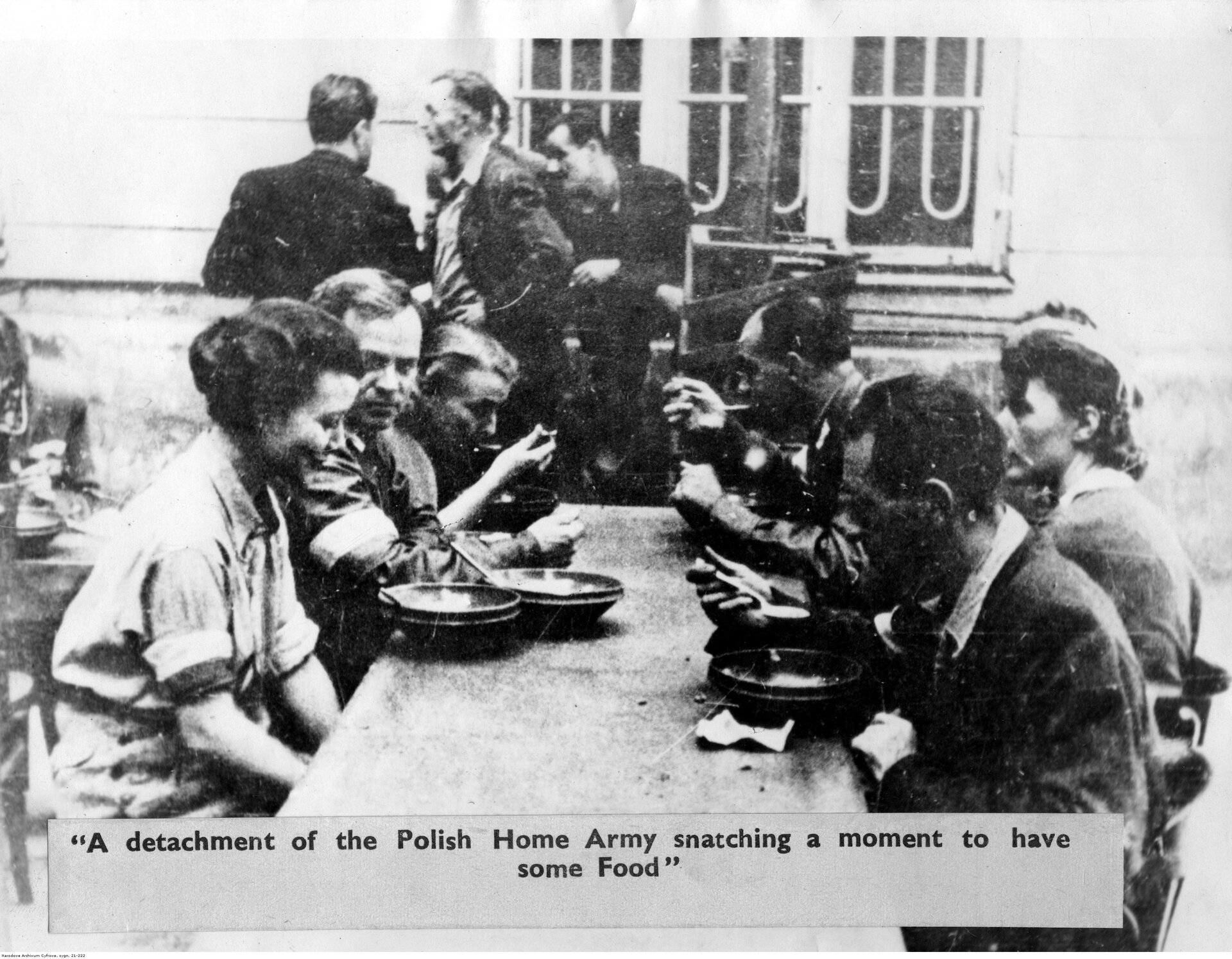 Zdjęcie przedstawia sześć osób siedzących przy stole. Są to kobiety i mężczyźni. Jedzą zupę. Na rękawach po lewej stronie mają szerokie, białe opaski. W tle ukazani są czterej mężczyźni stojący przy oknie budynku. Pod spodem znajduje się podpis: A detachment of the Polish Home Army snatching a moment to have some Food.