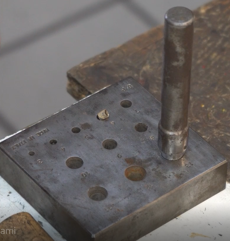 Ilustracja przedstawia kastownik. To metalowa blaszka w kształcie kwadratu. Na blaszce znajduje się jedenaście otworów, w ostatni największy otwór wbite jest narzędzie w kształcie walca z ostrym zakończeniem.