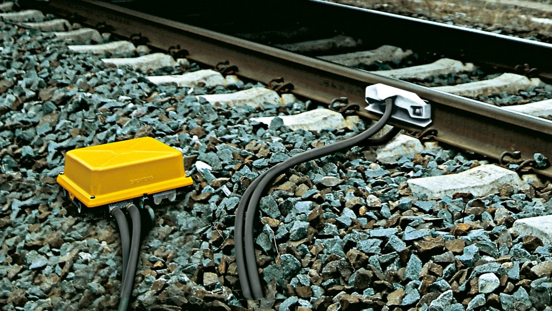 Ilustracja przedstawia jeden z odbiorników obwodów torowych i czujników torowych. Jest to niewielka żółta skrzynka umiejscowiona obok torów. Jest ona połączona przewodami z szynami. 