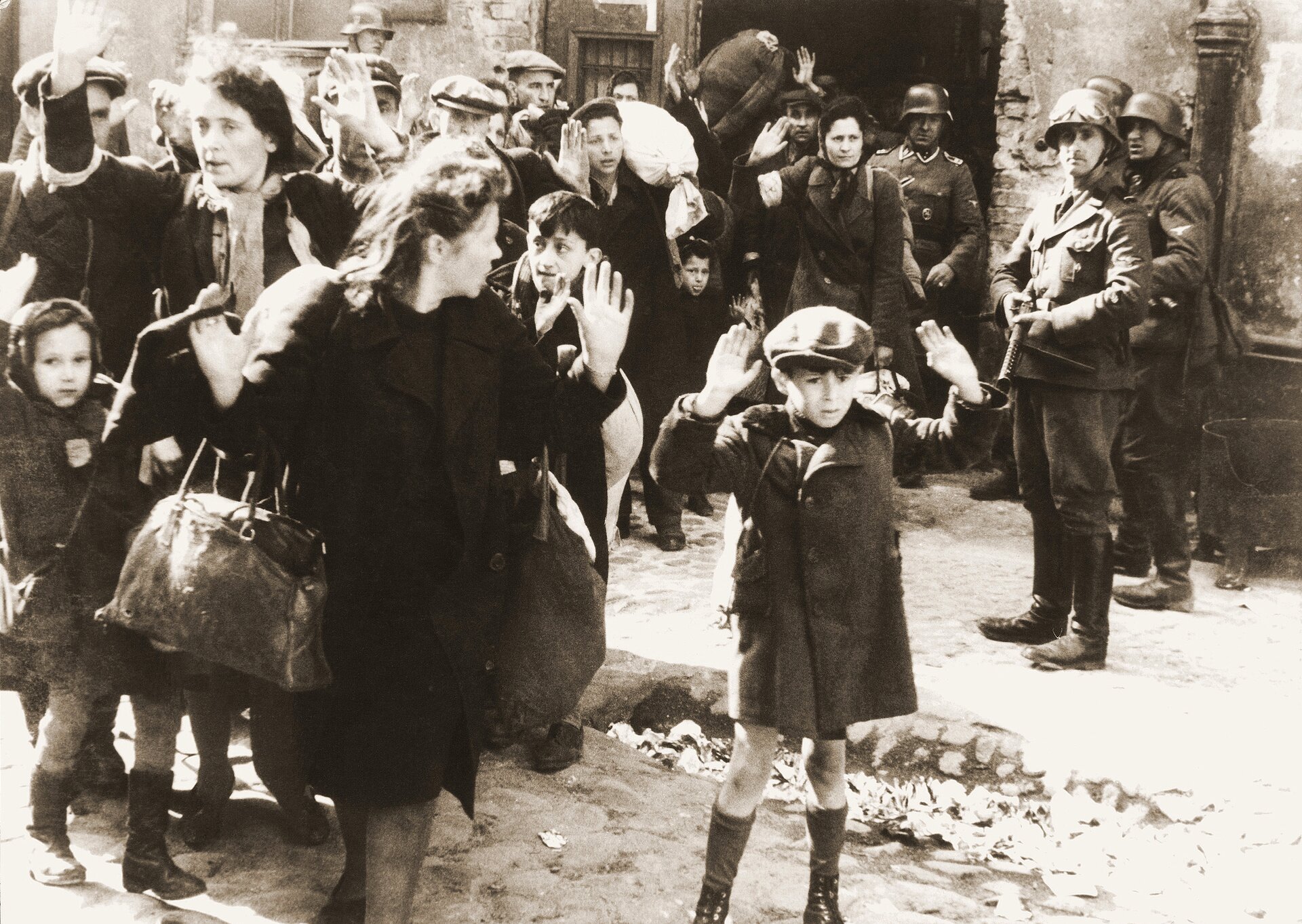 Z bramy budynku wychodzi grupa osób z podniesionymi dłońmi. W grupie są kobiety i dzieci. Obserwują ich uzbrojeni żołnierze niemieccy. Jeden z nich ma skierowaną broń w stronę chłopca.
