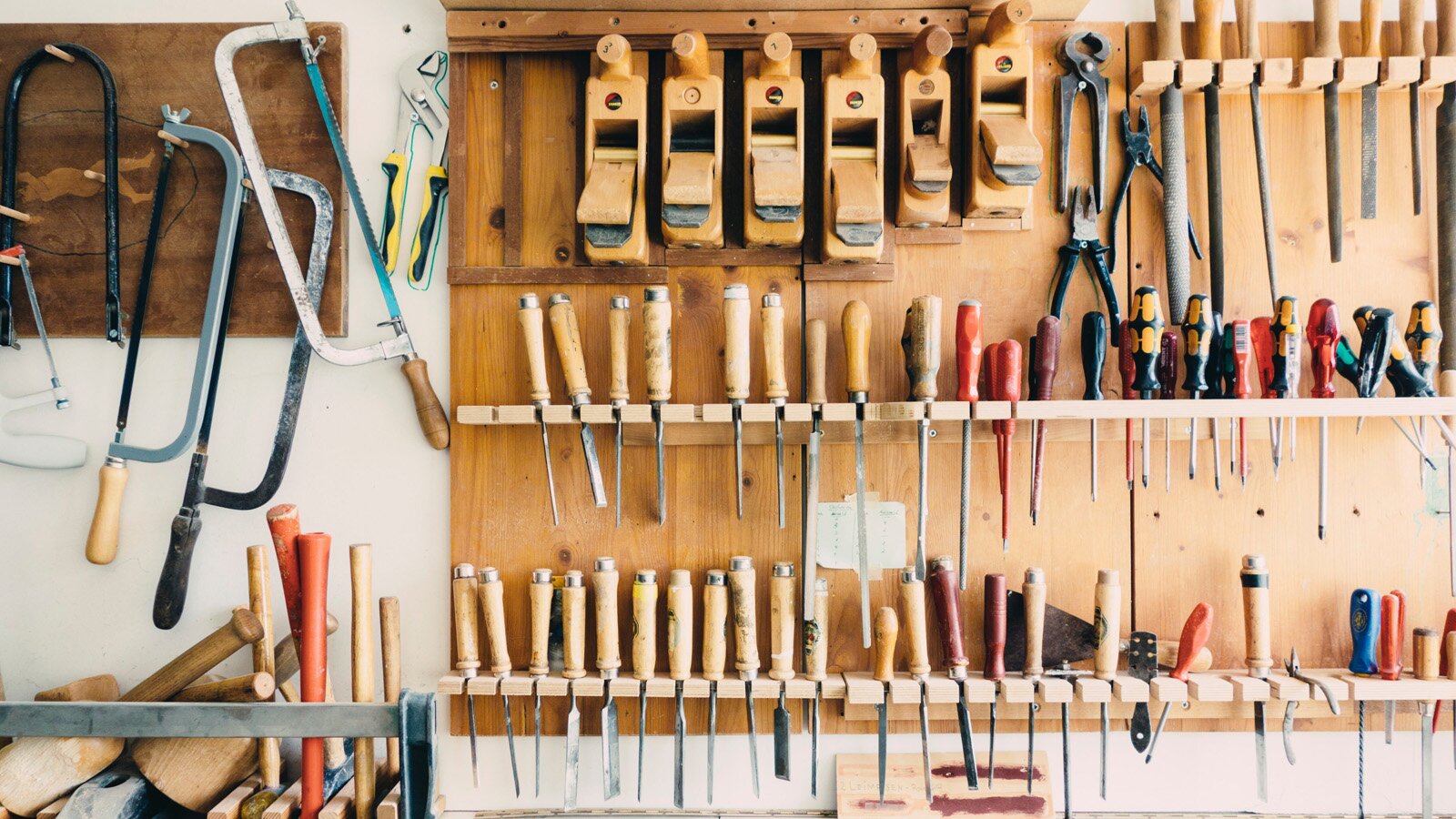Zdjęcie przedstawia ścianę, do której przymocowane są drewniane tablice z haczykami, półeczkami i zaczepami. Na większej tablicy zamocowane są różnego rozmiaru i rodzaju dłuta oraz heble, obcęgi i pilniki. Na mniejszej tablicy po lewej zawieszone są piły ręczne. Poniżej po lewej stronie widoczny jest stojak, z którego wystają różne trzonki oraz leżą dwa drewniane młotki.