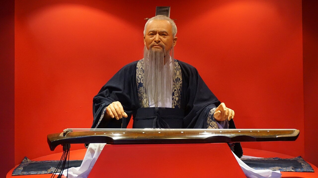 Kolorowe zdjęcie przedstawia mężczyznę o siwych włosach i długiej, siwej brodzie, który siedzi przy niskim stoliczku. Postać ubrana jest w kimono - tradycyjną japońską, długą szatę o szerokich rękawach, przewiązaną w pasie szeroką taśmą. Przed nim na stoliczku leży instrument szarpany. Mężczyzna unosi ręce nad nim. Tło jest jednolite, intensywnie czerwone.