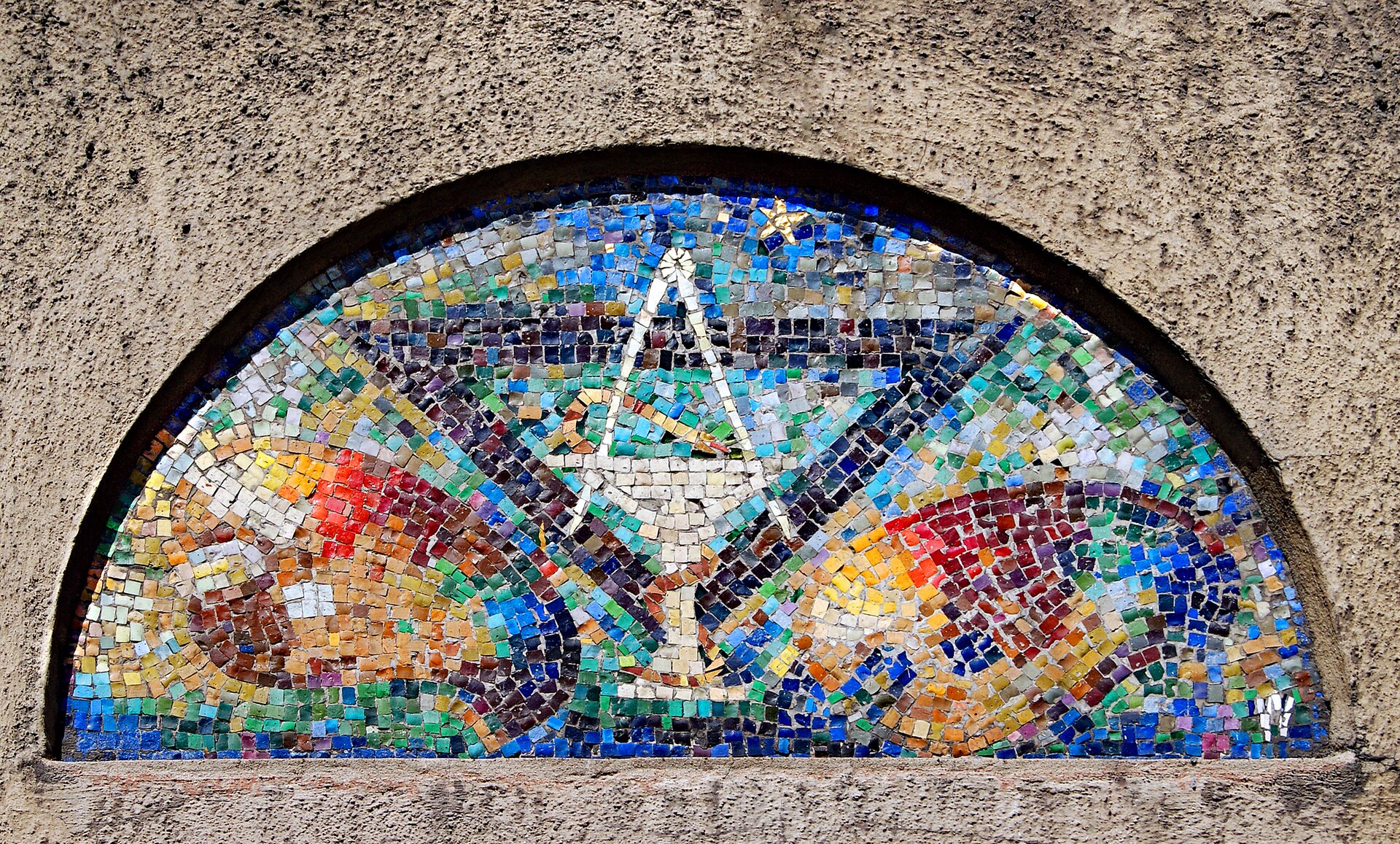 Fotografia przedstawia kolorową mozaikę umieszczoną na fragmencie ściany. Mozaika składa się z wielokolorowych miniaturowych kostek, które tworzą obraz - dwóch palet malarskich oraz pucharu przed którym  stoi figura kielicha z którego wystają dwa pędzle. Mozaika ma kształt półkola. 