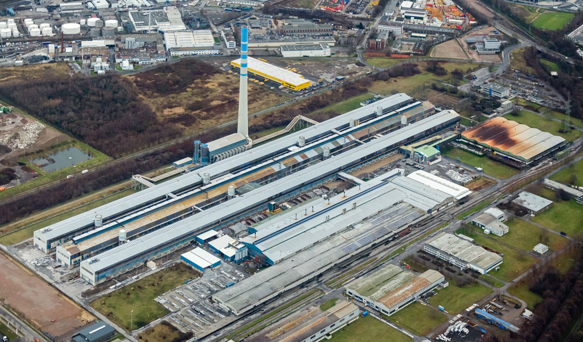 Zdjęcie huty aluminium oglądanej z lotu ptaku. Zakłady składają się z szeregu długich budynków. Jednemu z nich towarzyszy mniejszy kompleks z wysokim kominem. Widok innych budynków przemysłowych, mieszkalnych i magazynów, a także drogi prowadzącej do huty i samochodów na niej wskazują, że kompleks jest naprawdę wielki.