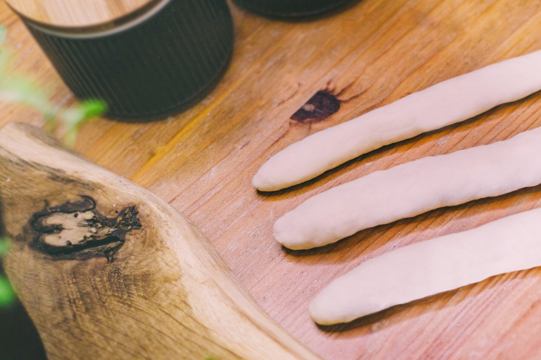 Zdjęcie przedstawia krok trzeci formowania precli. Zdjęcie wykonane jest od góry pod skosem. Widoczny jest ciemnobrązowy drewniany blat. W prawej części zdjęcia znajdują się trzy ułożone poziomo wałki ciasta. W górnej części grafiki dostrzec można fragmenty dwóch czarnych puszek z jasnobrązowymi wieczkami.