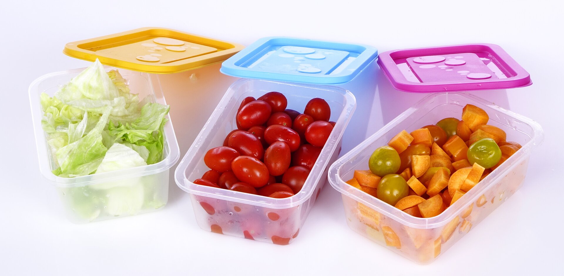 Fotografia przedstawia pokrojone surowe warzywa (sałatę, pomidory i marchew) umieszczone w plastikowych pojemnikach na żywność. Pojemniki mają prostokątny kształt i są otwarte. Kolorowe pokrywki (kolejno od lewej: żółta, niebieska i różowa) opierają się o krótszą krawędź pojemnika odsłaniając ich wnętrze. W pierwszym pojemniku od lewej strony znajduje się pokrojona sałata, w drugim czerwone pomidorki koktajlowe, a w trzecim pokrojona marchewka.