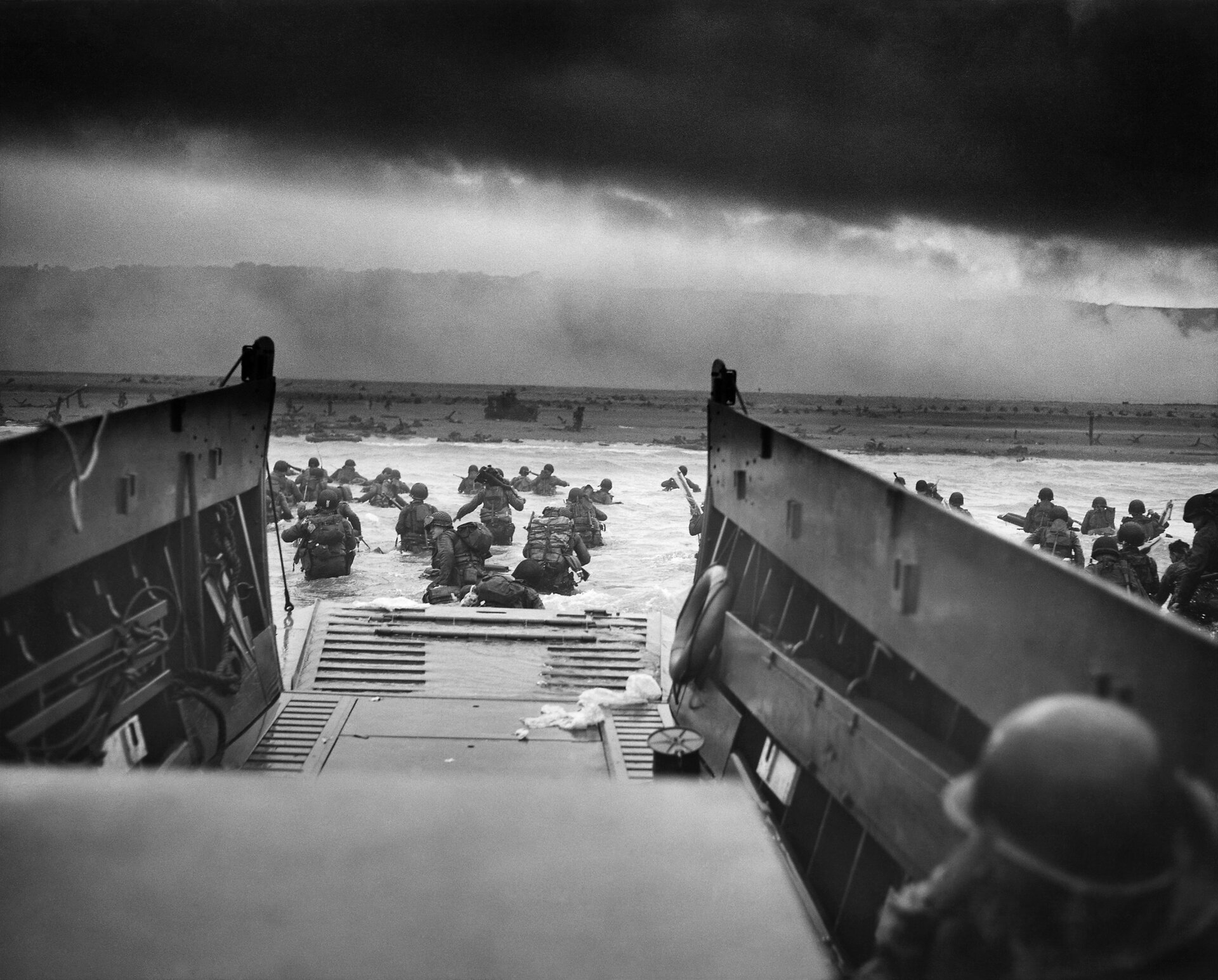Zdjęcie przedstawia plaże z perspektywy barki desantowej. Grupa żołnierzy czeka w barce. Inna grupa brodzi już w wodzie. W tle żołnierze z innych barek. Z wody wystają zapory przeciwczołgowe w postaci betonowych słupów. 