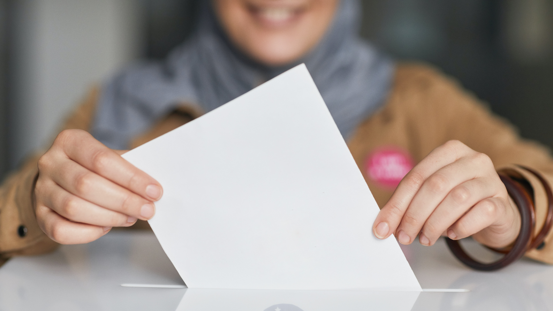 Zdjęcie przedstawia osobę wrzucającą kartkę do urny. Na pierwszym planie widoczne są dłonie osoby wsuwającej kartkę do urny. W tle widoczna jest rozmyta sylwetka tej osoby.