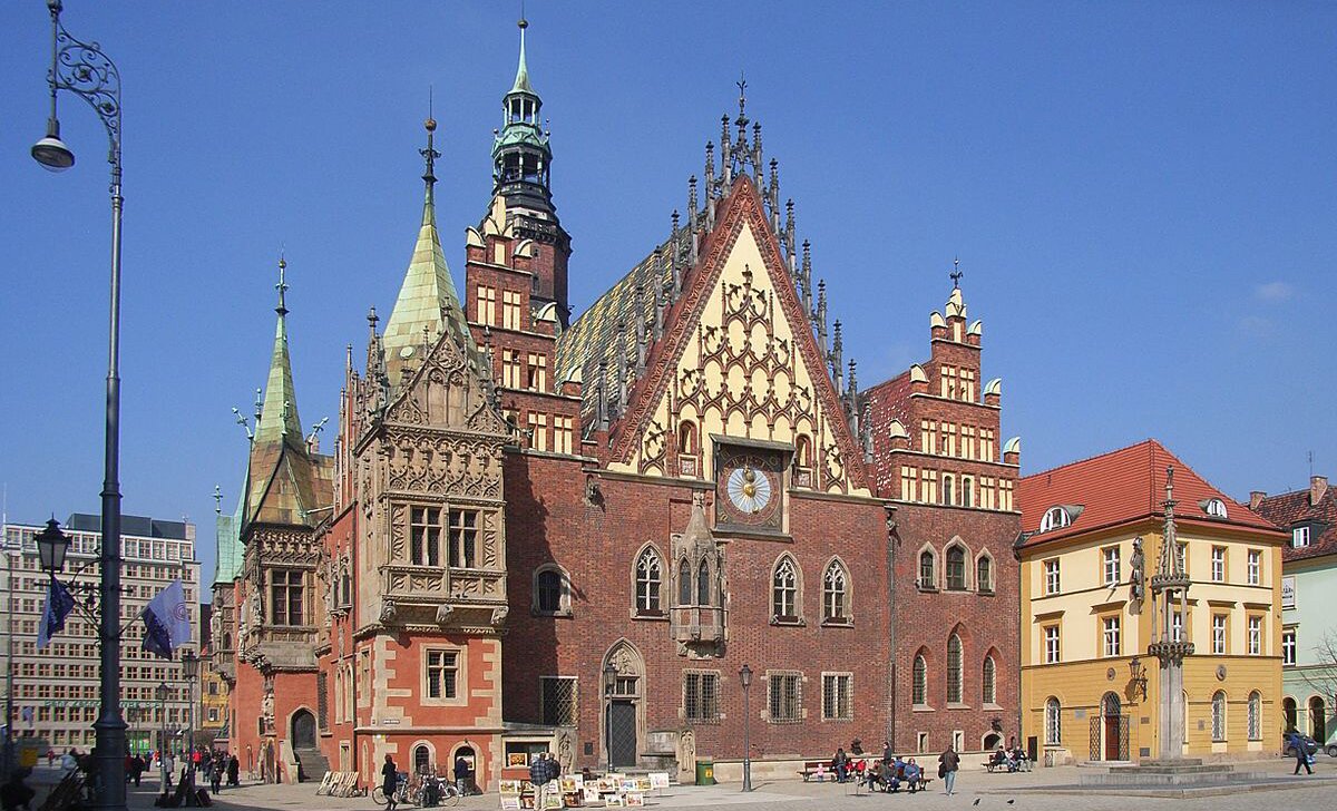 Fotografia barwna przedstawiająca wrocławski ratusz od strony południowo-wschodniej. Wschodnia, główna elewacja ratusza jest ceglana. Znajduje się na niej zegar z 1580 r. Widoczna część elewacji południowej pomalowana jest na rdzawoczerwono. Wystają z niej kamienne, bogato zdobione wykusze, czyli zwieszające się nad ziemią części budynku kryte osobnymi daszkami. Nad dwupiętrową budowlą góruje 66-metrowa wieża. Color photography depicting the Wroclaw Town Hall from the south-east side. The eastern, main elevation of the town hall is brick. There is a clock from 1580 on it. The visible part of the southern façade is painted in rust-red. There are stone, richly ornamented bay windows, which hang over the ground, parts of the building covered with separate roofs. A 66-meter tall tower dominates the two-story building.