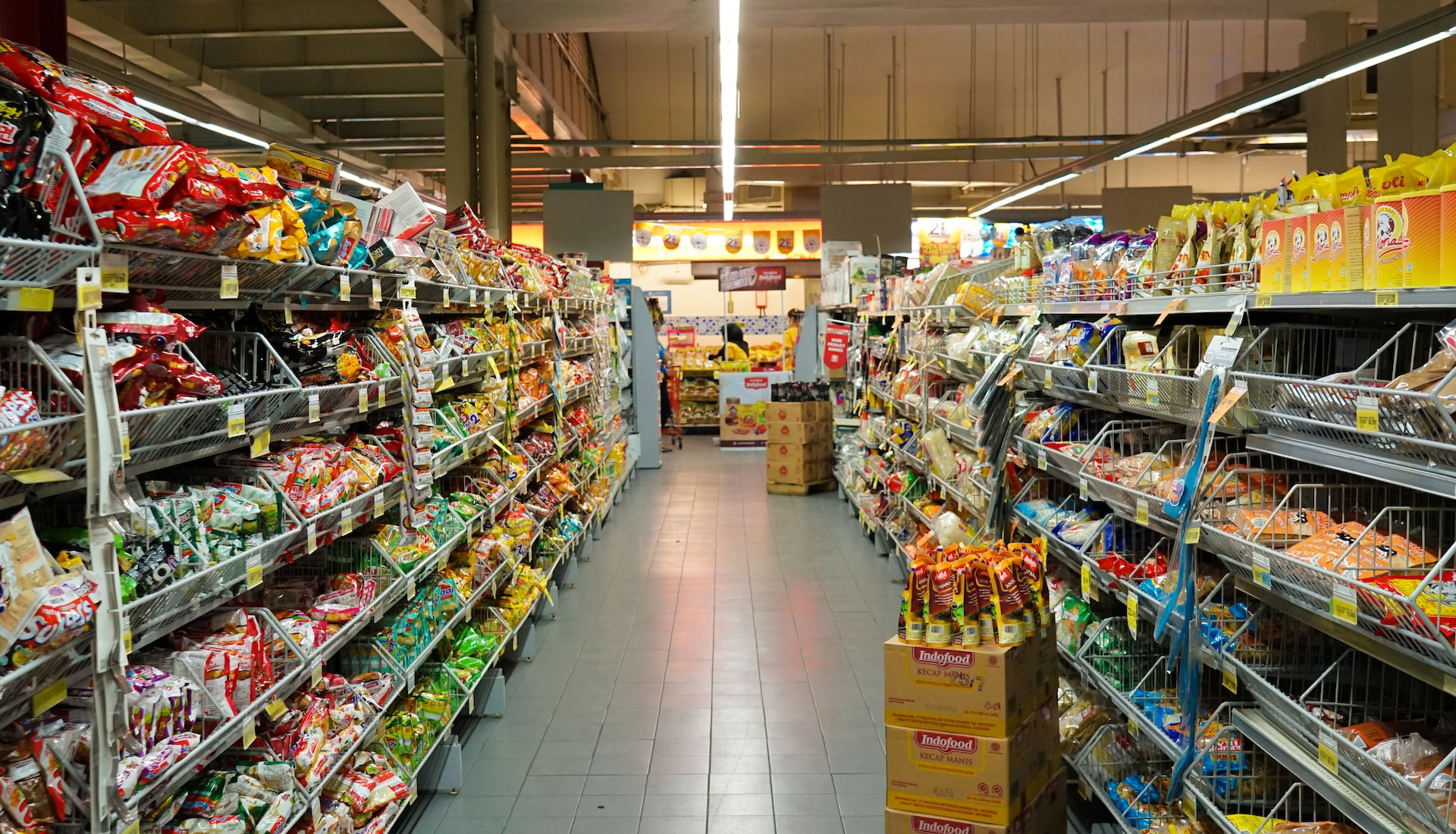 Zdjęcie przedstawia wnętrze sklepu samoobsługowego. Widoczna jest alejka sklepowa z półkami po lewej i prawej stronie, które są wypełnione różnymi opakowaniami ze słodyczami i przekąskami. Na końcu alejki widoczne jest oświetlone stoisko z pieczywem, przy którym stoją trzy osoby.