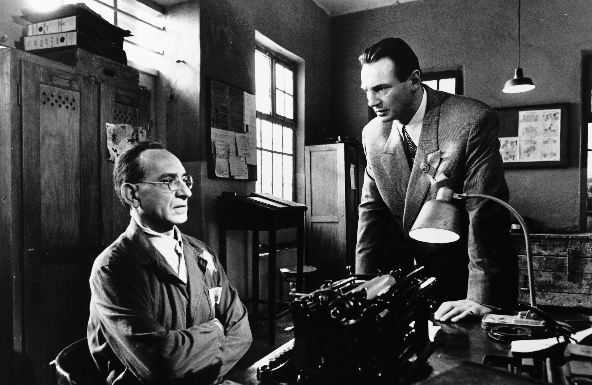 Ilustracja przedstawia kadr z filmu „Lista Schindlera” z widoczną dekoracją Ewy Braun z 1993 roku. Na ilustracji znajduje się dwóch mężczyzn w pomieszczeniu. Mężczyzna po lewej stronie siedzi na krześle przy biurku. Ma on krótkie włosy zaczesane do tyłu i duże zakola. Jego okrycie wierzchnie jest z długim rękawem. Na nosie ma okrągłe okulary. Jego ręce są skrzyżowane, a dłonie ukryte pod ramionami. Przed nim na biurku stoi maszyna do pisania, a dalej lampka. Za nim znajduje się dwudrzwiowa szafa, a na niej leżą segregatory. Nad szafą jest okno. Drugi mężczyzna opiera się dłońmi o biurko. Ma krótkie włosy zaczesane do tyłu. Ubrany jest w marynarkę, białą koszulę i wzorzysty krawat. Na klapie marynarki ma przypięty okrągły znaczek i białą poszetkę w kieszonce. Za nim znajduje się skrzynia, okno oraz na ścianie wisi tablica z przyczepionymi kartkami. Z sufitu zwisa lampa. W rogu pomieszczenia stoi szafa. Obok niej jest okno, wysokie biurko i okrągłe krzesło. Nad biurkiem wisi tablica z przyczepionymi kartkami. Kolorystyka ilustracji jest biało‑czarna. 