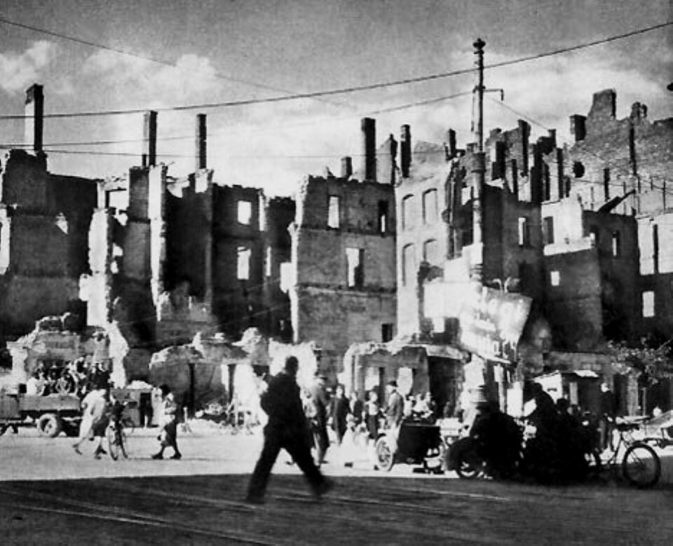 Zdjęcie przedstawia ruiny Warszawy po wyzwoleniu. Środkiem drogi przechodzi mężczyzna, za nim widać ludzi przemieszczających się pieszo, rowerami i na wozach. W tle sterczą kominy i ściany  zrujnowanych domów.