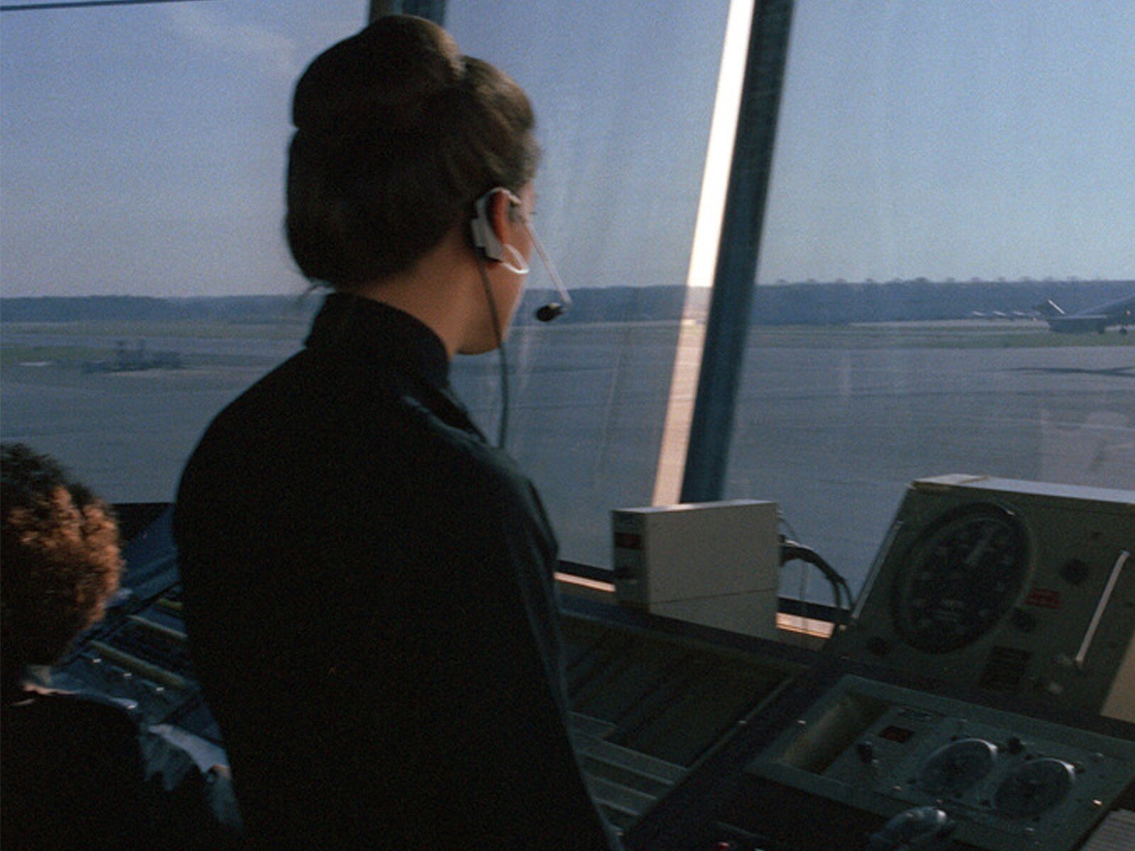 Zdjęcie przedstawia młodą kobietę, w ujęciu od pasa w górę, stojącą w wieży kontrolnej lotniska. Kobieta jest zwrócona w prawo i patrzy przez duże okna na płytę lotniska. Jest ubrana w ciemną koszulę. Na głowie ma słuchawki z mikrofonem ustawionym przy ustach. Przed nią widoczna jest aparatura. Przez okna wieży kontrolnej widoczna jest płyta lotniska i ogon samolotu stojącego po prawej stronie.