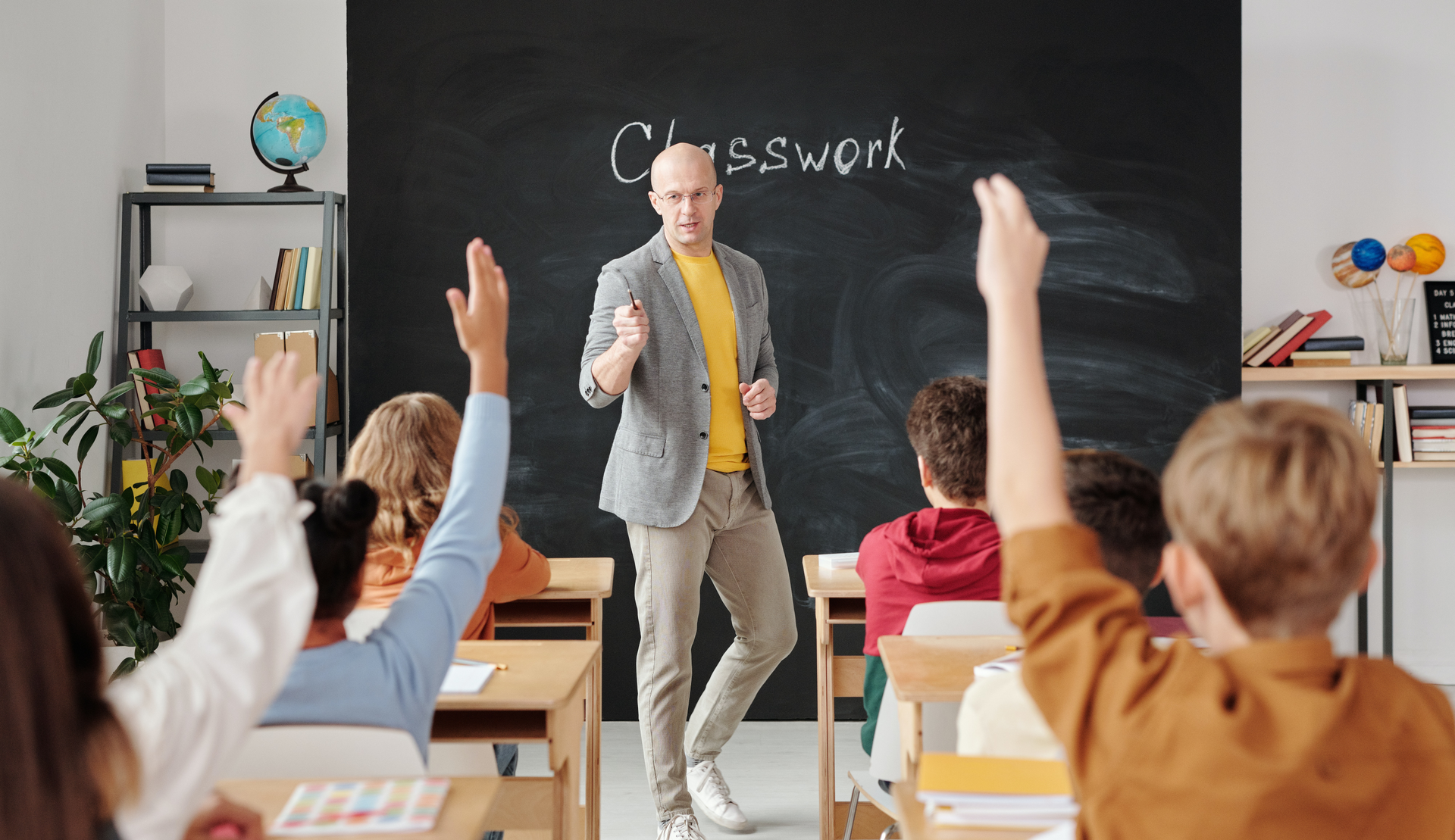 Zdjęcie przedstawia klasę szkolną. Na środku, przodem do kadru stoi łysy nauczyciel w okularach. W ławkach, tyłem do kadru siedzą uczniowie. Podnoszą ręce do odpowiedzi. W tle widoczna jest tablica z napisem: "Classwork".