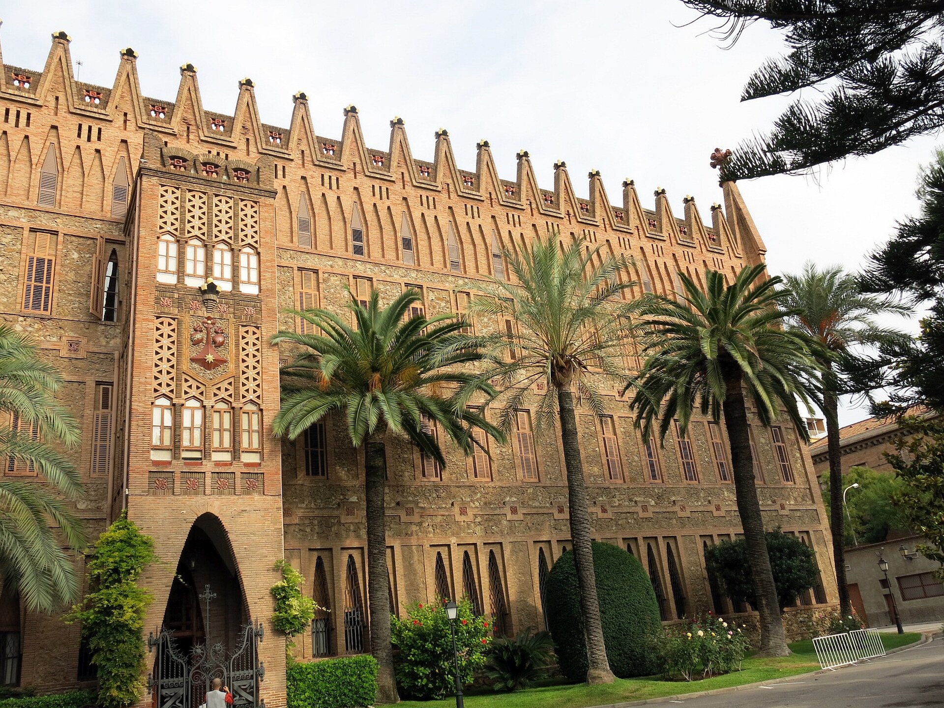 Ilustracja interaktywna o kształcie poziomego prostokąta przedstawia fotografię Kolegium Teresjańskie w Barcelonie zaprojektowanego przez Antonio Gaudiego. Budowla zbudowana jest z cegły oraz posiada elementy ceramiki. Kształt budynku jest prostokątny, cały przyozdobiony ceglanymi pinaklami oraz ukoronowany krzyżem Gaudiego i herbami przedstawiającymi figury Zakonu Terezjańskiego. Przed budowlą znajduje się trawnik z krzewami i palmami. 