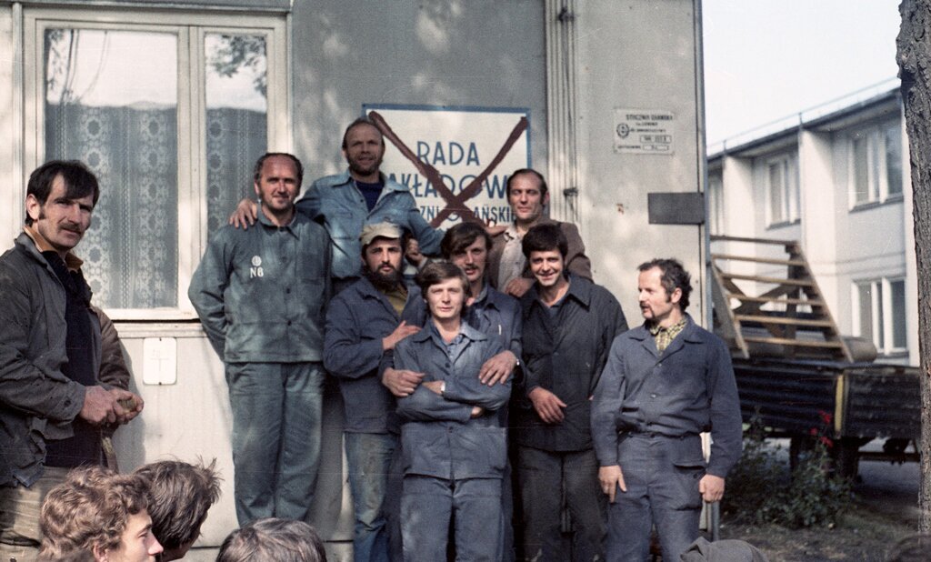 Zdjęcie przedstawia dwunastu pracowników Stoczni. Są to osoby strajkujące. W większości mają ubrane granatowe stroje robocze. Ośmioro spośród robotników zakrywa przekreśloną tablicę Rady Zakładowej, przymocowaną do ściany. W tle zdjęcia znajduje się przyczepa z drabiną oraz niskie zabudowania. W oknach budynków są firany.