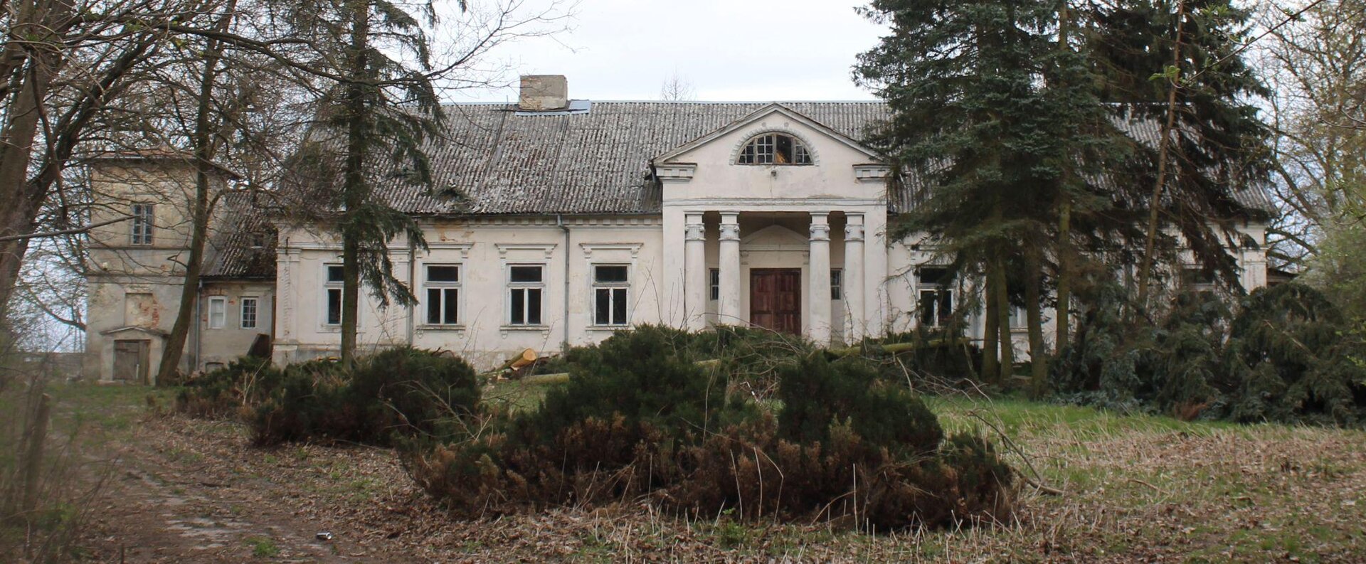 Zdjęcie przedstawia biały dworek z szarym, skośnym dachem. Część budynku, znajdująca się nad wejściem, jest wysunięta do przodu i wsparta na czterech kolumnach. Wokół dworku rosną zielone choinki i bezlistne drzewa. Przed budynkiem rosną niskie, zielone krzewy, a także zielona trawa, częściowo przykryta liśćmi. Niebo jest błękitne, bezchmurne. Zdjęcie zostało wykonane w świetle dziennym.