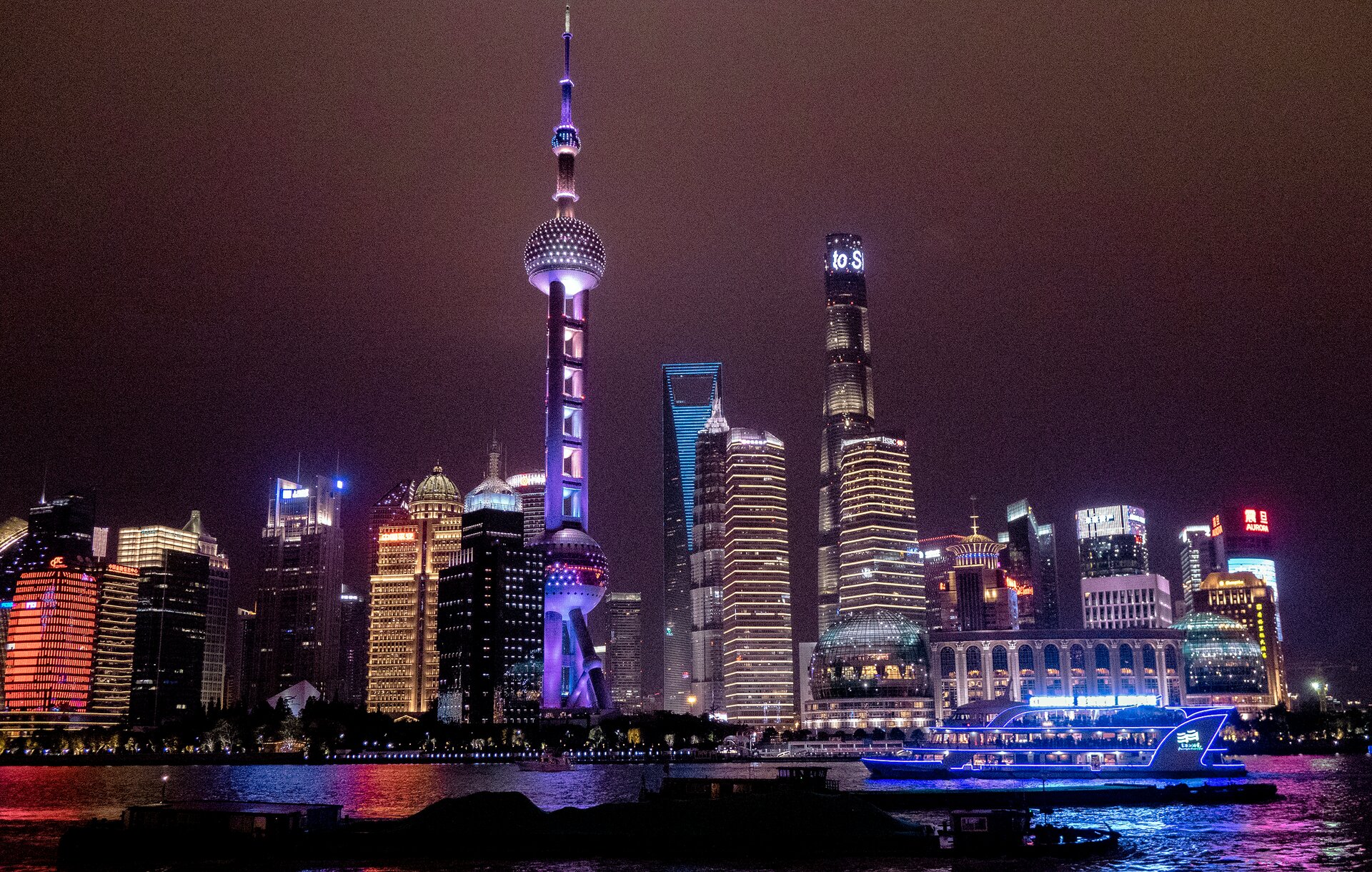 Fotografia przedstawia panoramę miasta. Na pierwszym planie widoczny jest zbiornik wodny z przepływającym, oświetlonym na niebiesko statkiem. Przy linii brzegowej widoczne są ciemne drzewa, a za nimi oświetlone budynki. W centrum nad nimi znajduje się budynek, którego konstrukcja składa się z dwóch dużych kul znajdujących się w dużej odległości od siebie, jedna nad drugą, podtrzymywanych przez grube filary zwieńczone iglicą. Jest podświetlony na różowo‑fioletowo. Inne widoczne budynki to między innymi: po prawej stronie znajdują się ciemne wieżowce oświetlone na złoto, po lewej stronie znajdują się masywny budynek z czerwonymi światłami oraz wysokie, jasne kolumny. Tło stanowi ciemne niebo.