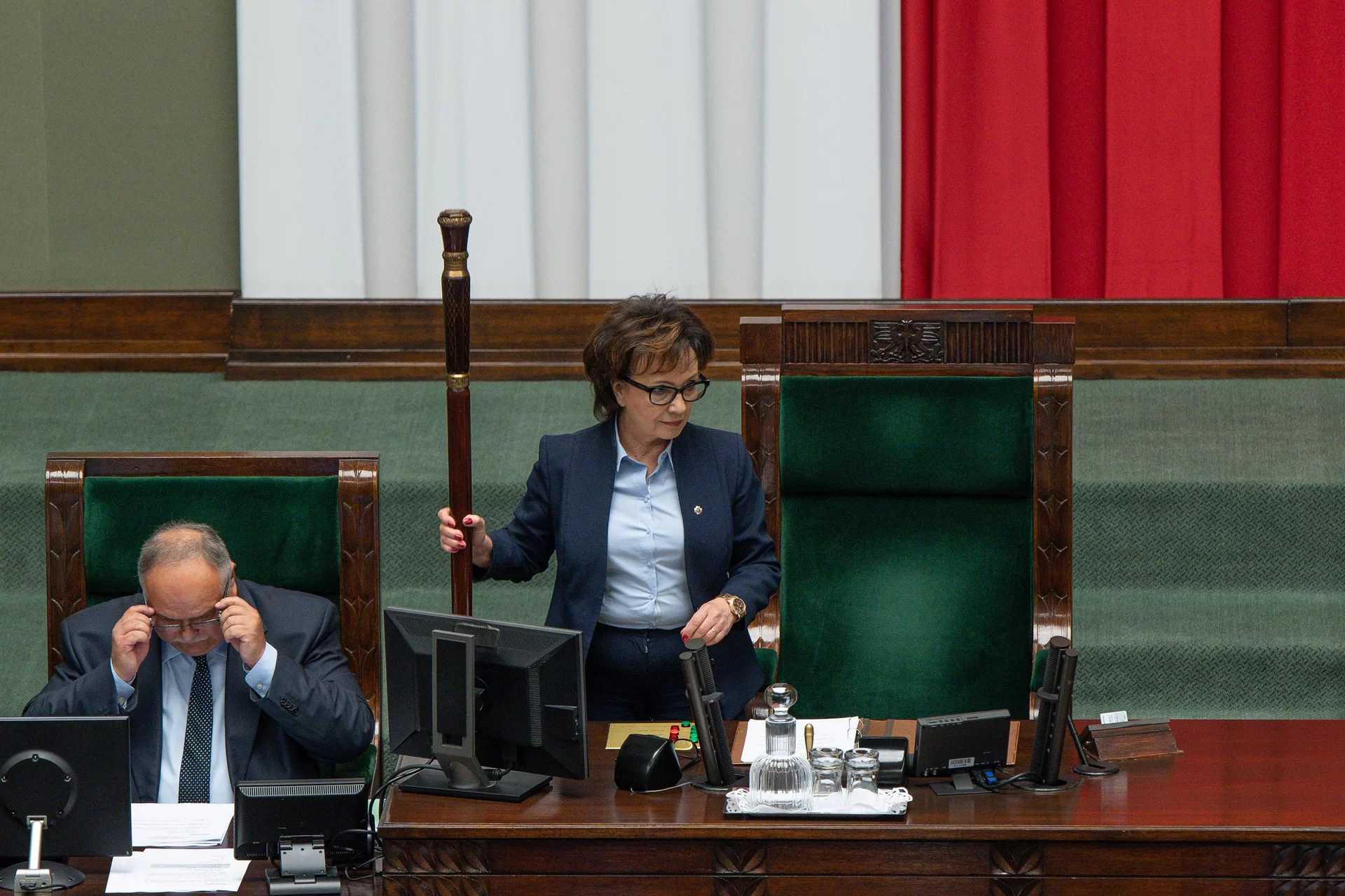 Zdjęcie przedstawia marszałkinię Sejmu, która trzyma w ręku laskę marszałkowską. Kobieta ma ciemną, krótką, nachodzącą na czoło fryzurę oraz okulary w grubych, czarnych oprawkach. Ubrana jest w ciemnoniebieską garsonkę. Stoi przed sejmową ławą, na której znajdują się komputer, mikrofony oraz karafka z wodą.