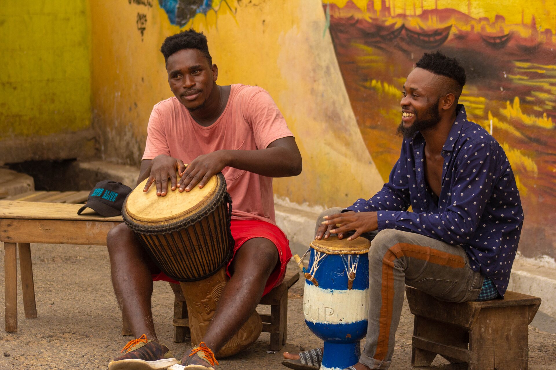 Zdjęcie przedstawia dwóch czarnoskórych mężczyzn grających na bębnach. Znajdują się na zewnątrz przed budynkiem, na którego ścianie znajduje się malowidło łodzi na wodzie. Mężczyźni siedzą na drewnianych ławkach. Jeden z nich ma na sobie różową koszulkę z krótkim rękawem, krótkie, czerwone spodenki oraz brązowo zielone buty sportowe z pomarańczowymi sznurówkami. Bęben, na którym gra jest brązowy i średniego rozmiaru. Drugi mężczyzna ma na sobie odpiętą, niebieską koszulę z długim rękawem w białe kropki, szare jeansy z pomarańczowymi lampasami oraz niebiesko czarne klapki. Gra na małym bębnie, koloru biało niebieskiego.