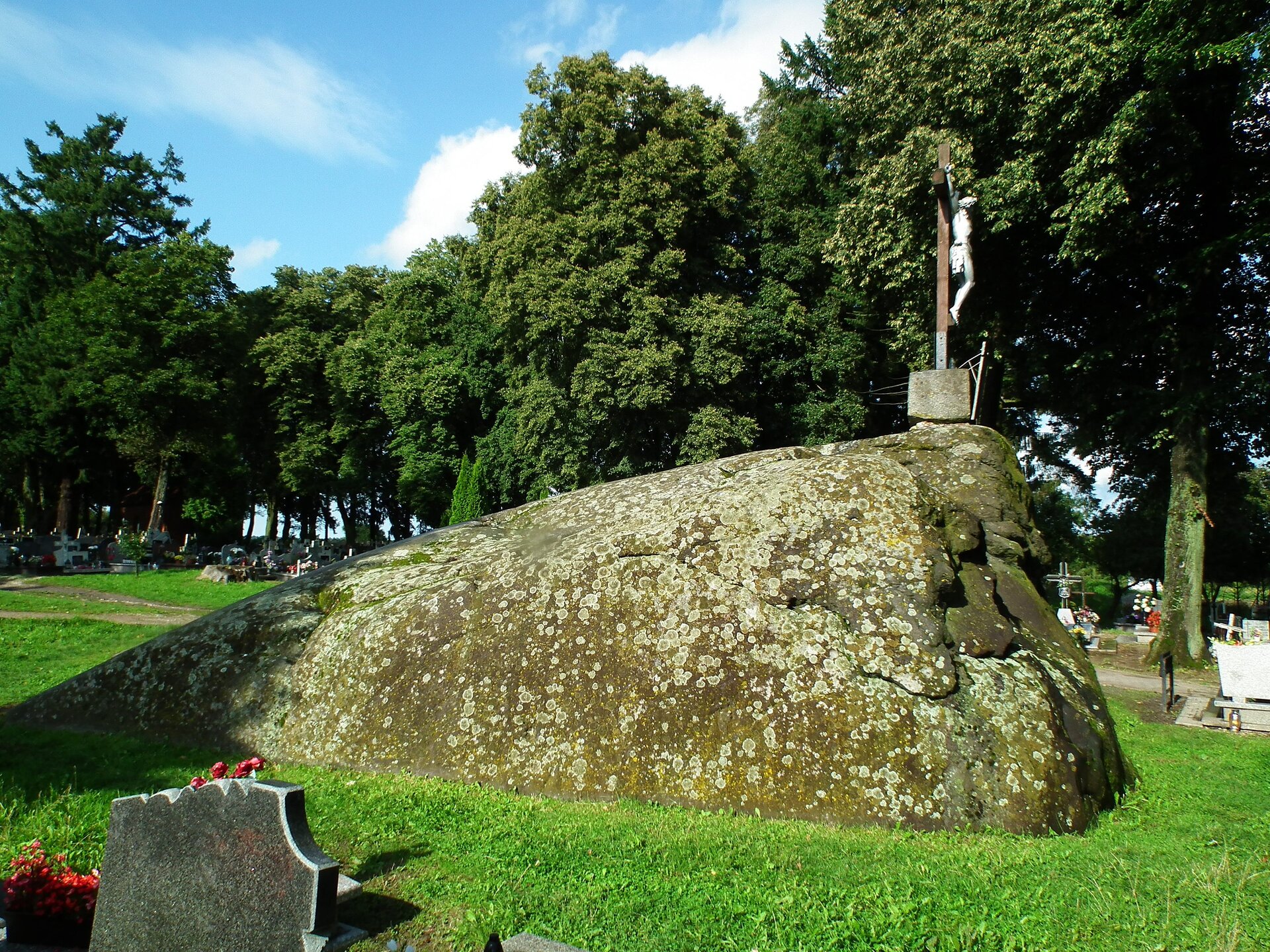 Zdjęcie przedstawia głaz narzutowy. Jeden bok jest wysoki i opada do samego dołu. Na szczycie znajduje się figura Jezusa na krzyżu. Wokół znajdują się nagrobki oraz drzewa.