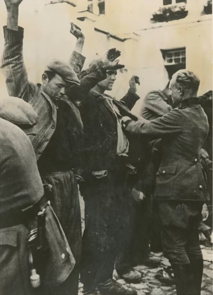 Kliknij, aby powiększyć Zdjęcie przedstawia kilku mężczyzn stojących w szeregu. Ubrani są w marynarki i czapki. Mają ręce uniesione do góry. Niemiecki żołnierz przeszukuje jednemu z nich wewnętrzną kieszeń marynarki. W tle kamienica.