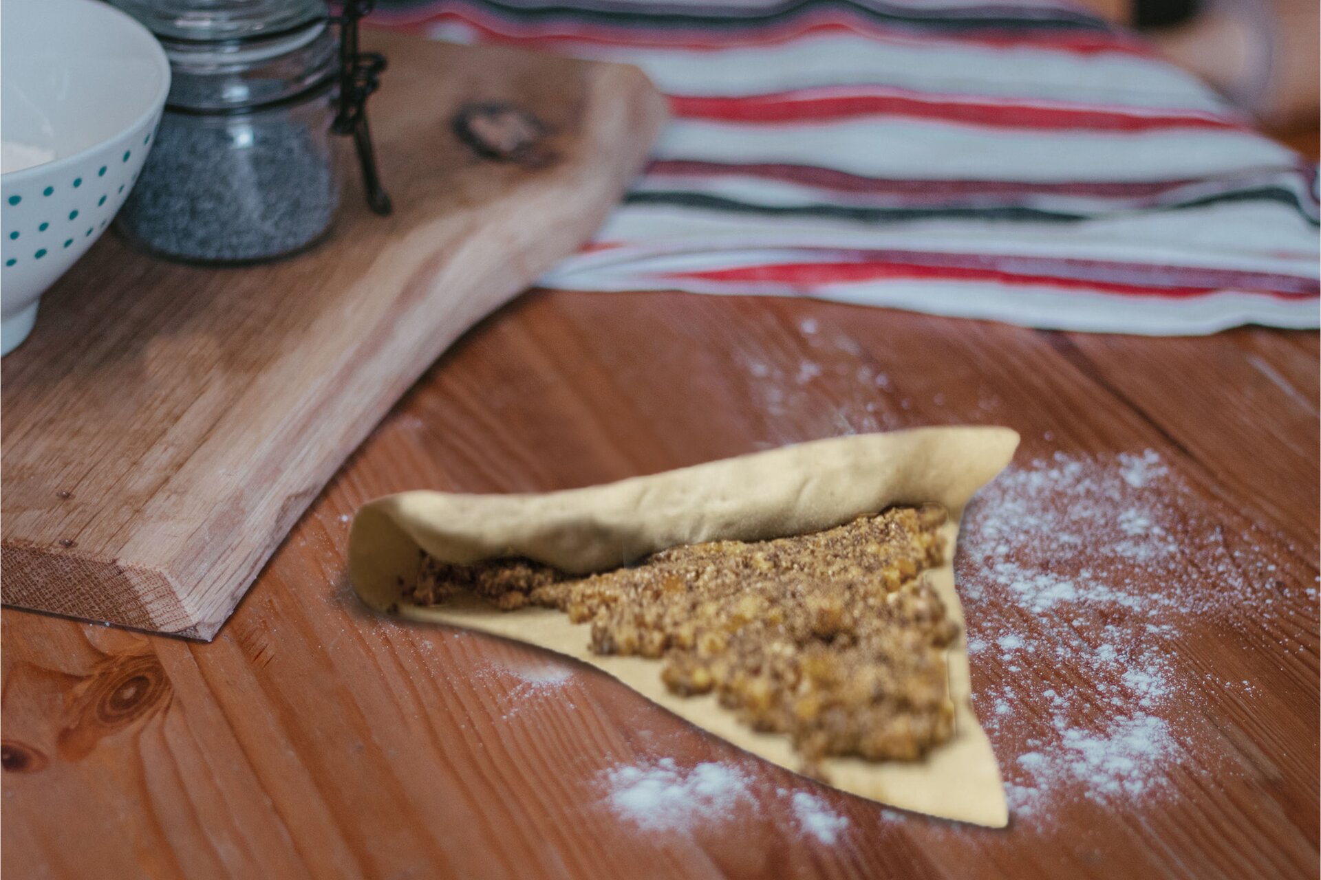 Zdjęcie przedstawia krok czwarty, piąty i szósty formowania rogali świętomarcińskich. Ujęcie wykonane jest od góry pod skosem. Widoczny jest ciemnobrązowy drewniany blat na którym w środkowej części grafiki znajduje się trójkątny płaski płat ciasta. Na całym płacie rozsmarowana jest masa będąca nadzieniem. Płat ciasta został zwinięty od podstawy trójkąta. Dookoła ciasta znajduje się rozsypana biała mąka. W lewej górnej części zdjęcia widoczna jest jasnobrązowa drewniana deska ułożona pionowo. Na desce stoi biała miseczka w niebieskie małe kropki. Nad nią dostrzec można przezroczysty słoik z czarnym klipsem w środku z widocznym niebieskim makiem. W górnej części zdjęcia widoczna jest biała ścierka w czerwone i czarne paski ułożona poziomo. Lewa część ścierki znajduje się pod drewnianą deską.