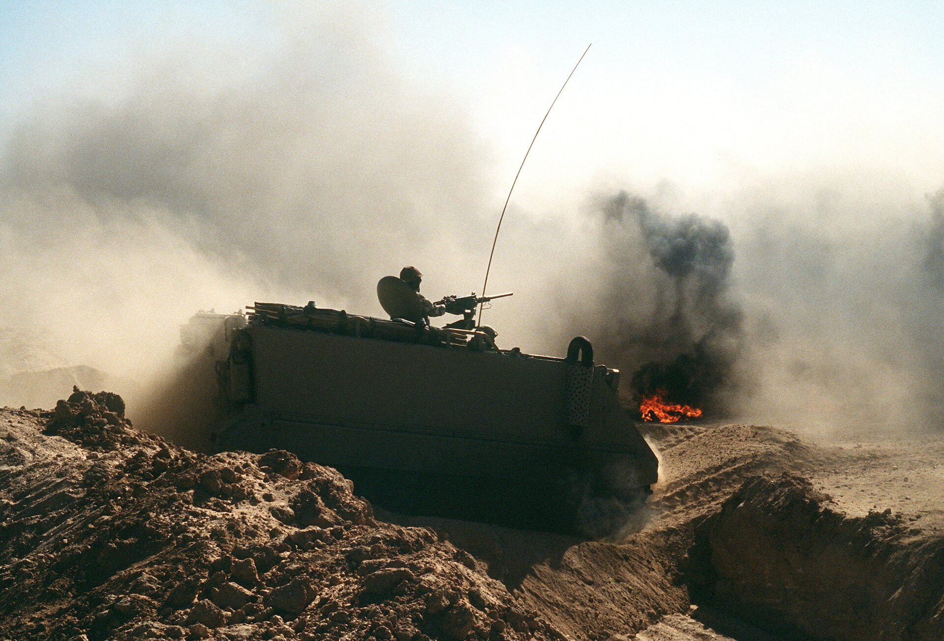 Zdjęcie przedstawia jałowy, piaszczysty teren. Pośrodku czołg, a na nim żołnierz celujący z karabinu maszynowego. Na drugim planie płomienie i chmura dymu.
