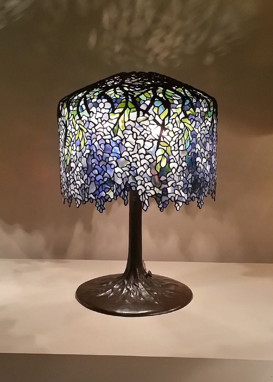 Fotografia przedstawia Lampę witrażową autorstwa Louisa Comfort Tiffanyego. Lampa stoi na szarym stole, ma okrągłą podstawkę, brązowy pałąk oraz witrażowy kolorowy klosz, na którym dominują barwy jasne oraz zielone. W górnej części lampa ma czarne zdobienia.