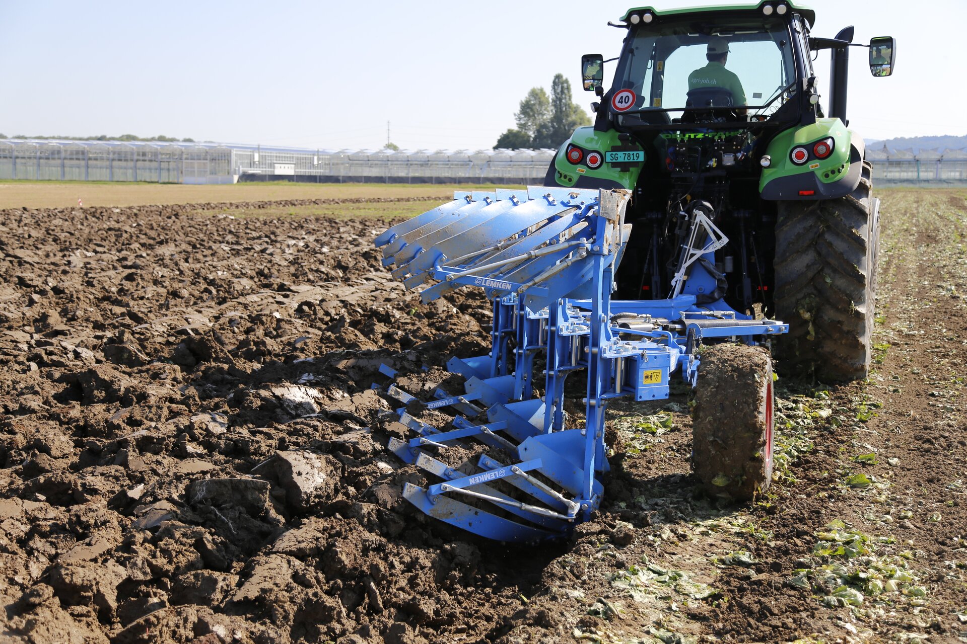 Zdjęcie pierwsze, podpisane traktor z urządzeniem rolniczym. Przedstawia traktor jadący po polu. Z tyłu przymocowany jest pług. W tle widoczne są zabudowania.