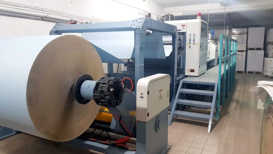 Zdjęcie przedstawia przekrawacz, w ujęciu z boku. Od lewej widoczna jest rola papieru, nawinięta na cylinder przymocowany za pomocą ramienia do podstawy. Dalej widoczna jest metalowa konstrukcja z czterech pionowych stelaży połączonych poziomymi poprzeczkami u góry, przez którą przechodzi rozwinięta taśma papieru. W tyle zdjęcia widoczne są 4 schody prowadzące na podest, na którym widoczny jest fragment prostopadłościennego elementu maszyny, najprawdopodobniej panelu sterującego.