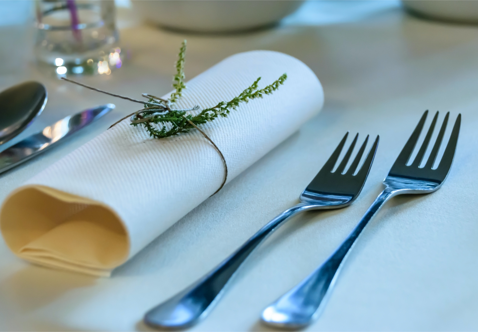 Zdjęcie przedstawia sztućce leżące na stole na obrusie. Po prawej znajdują się dwa widelce, mniejszy i większy. Pośrodku leży serwetka zwinięta w rulon, przewiązana sznurkiem i ozdobiona kawałkiem rośliny. Po lewej widoczny jest fragment noża i łyżki.